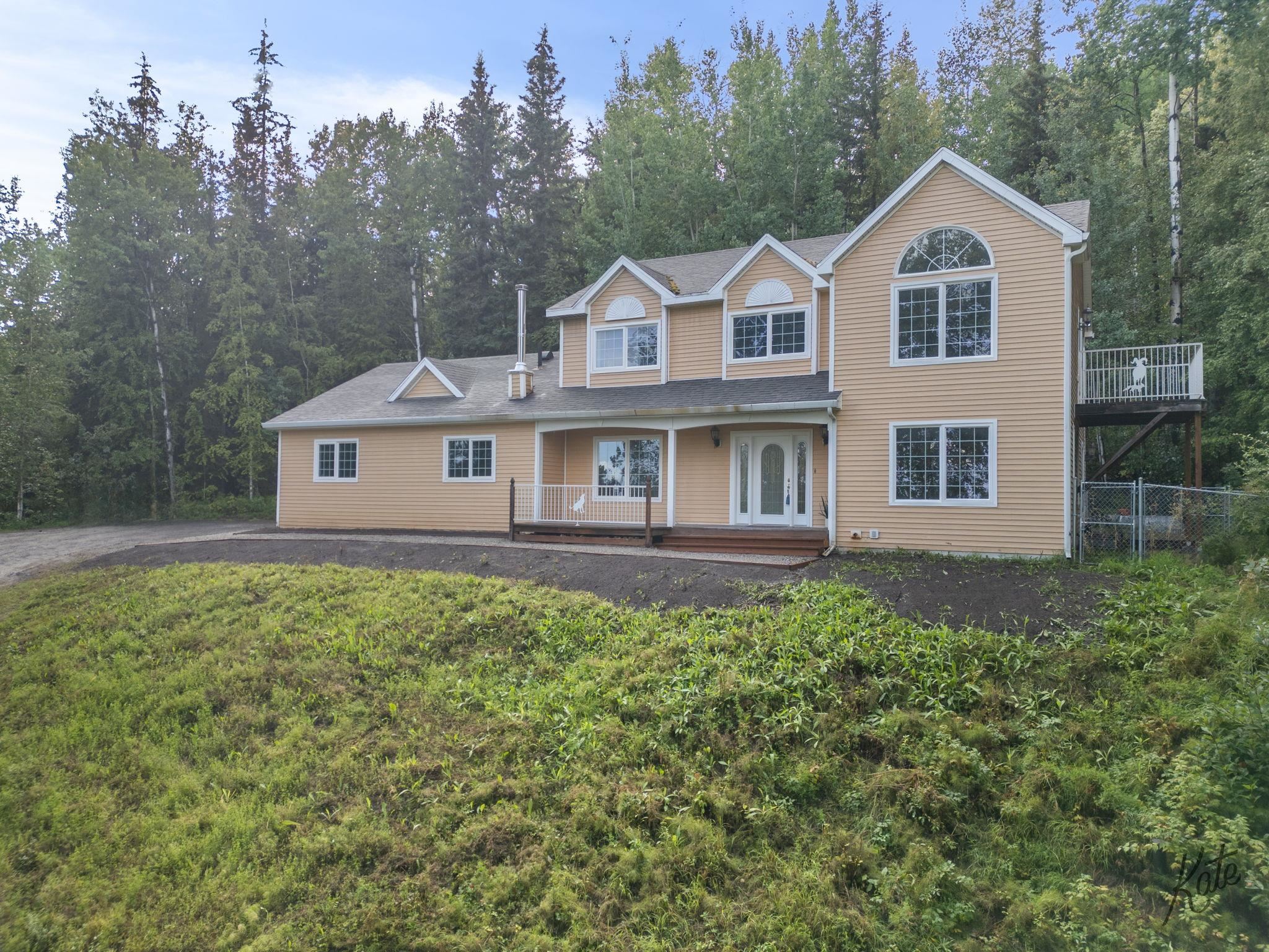 Tan two-story house with white-trimmed gables, a covered porch, and a raised deck beside woods.