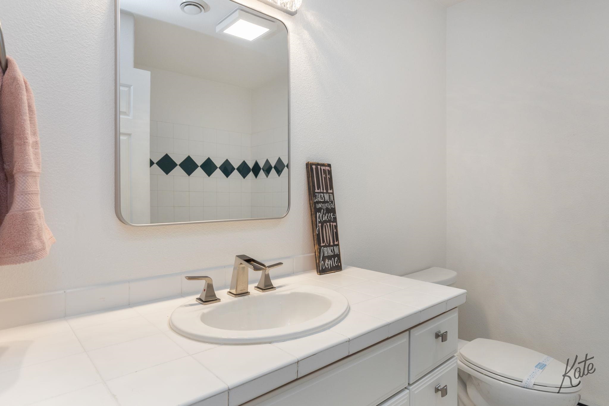 Bathroom vanity with a large mirror, white tiled counter, and dark diamond-pattern tile border.