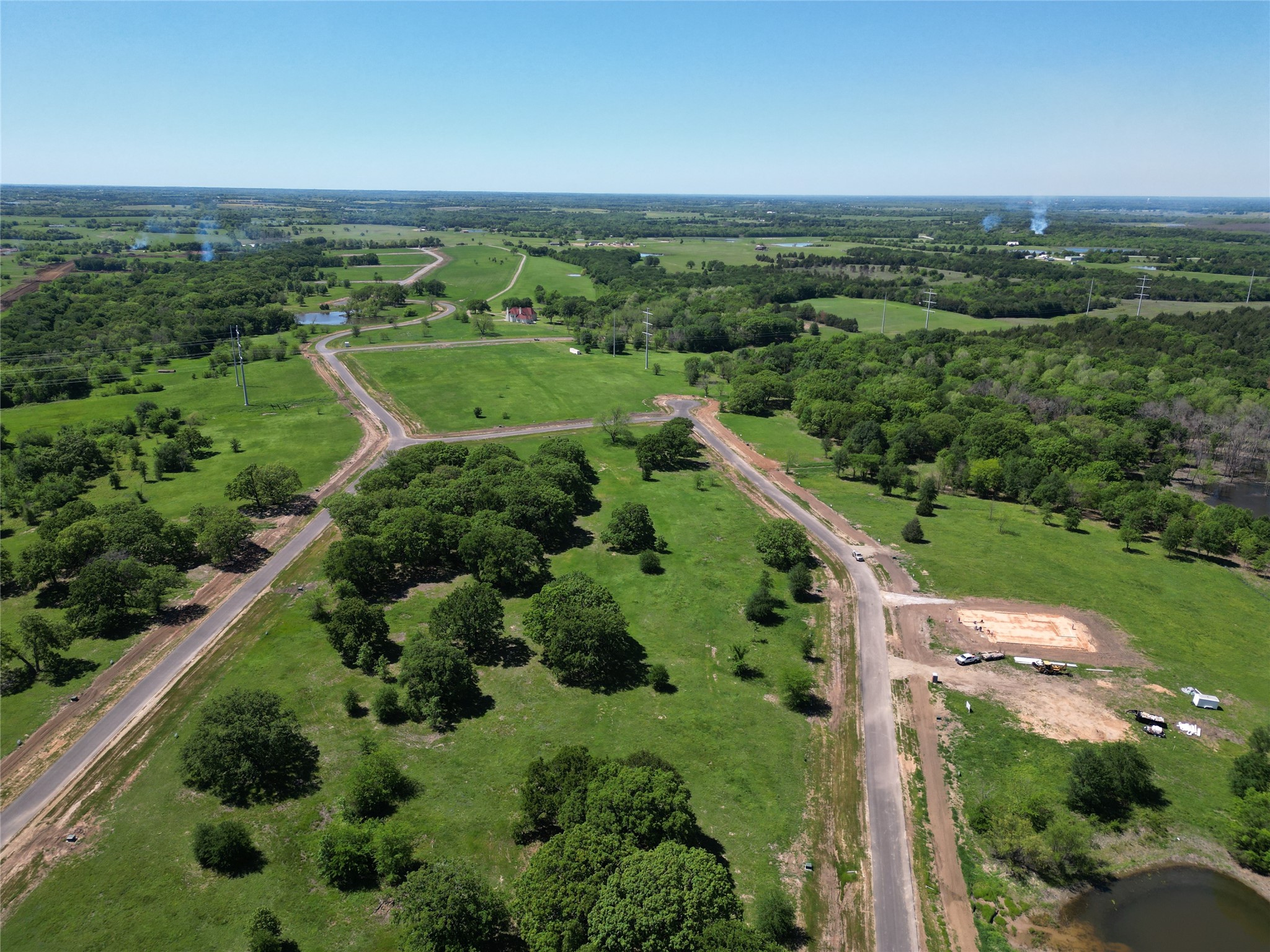 Aerial view of green acreage with winding roads, mature trees, and a home foundation under construction.