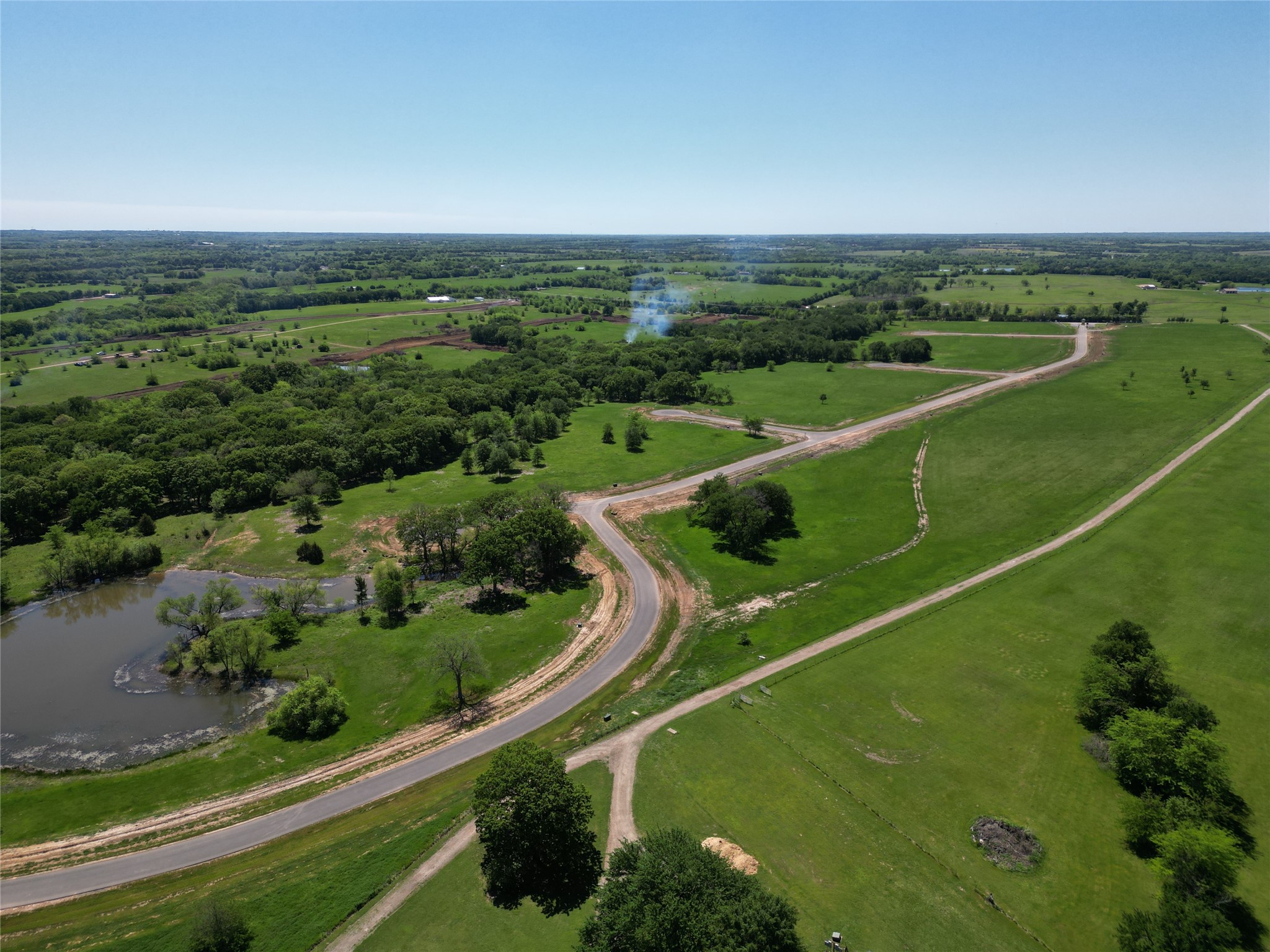 Aerial view of green lots with a winding paved road, mature trees, and a pond.