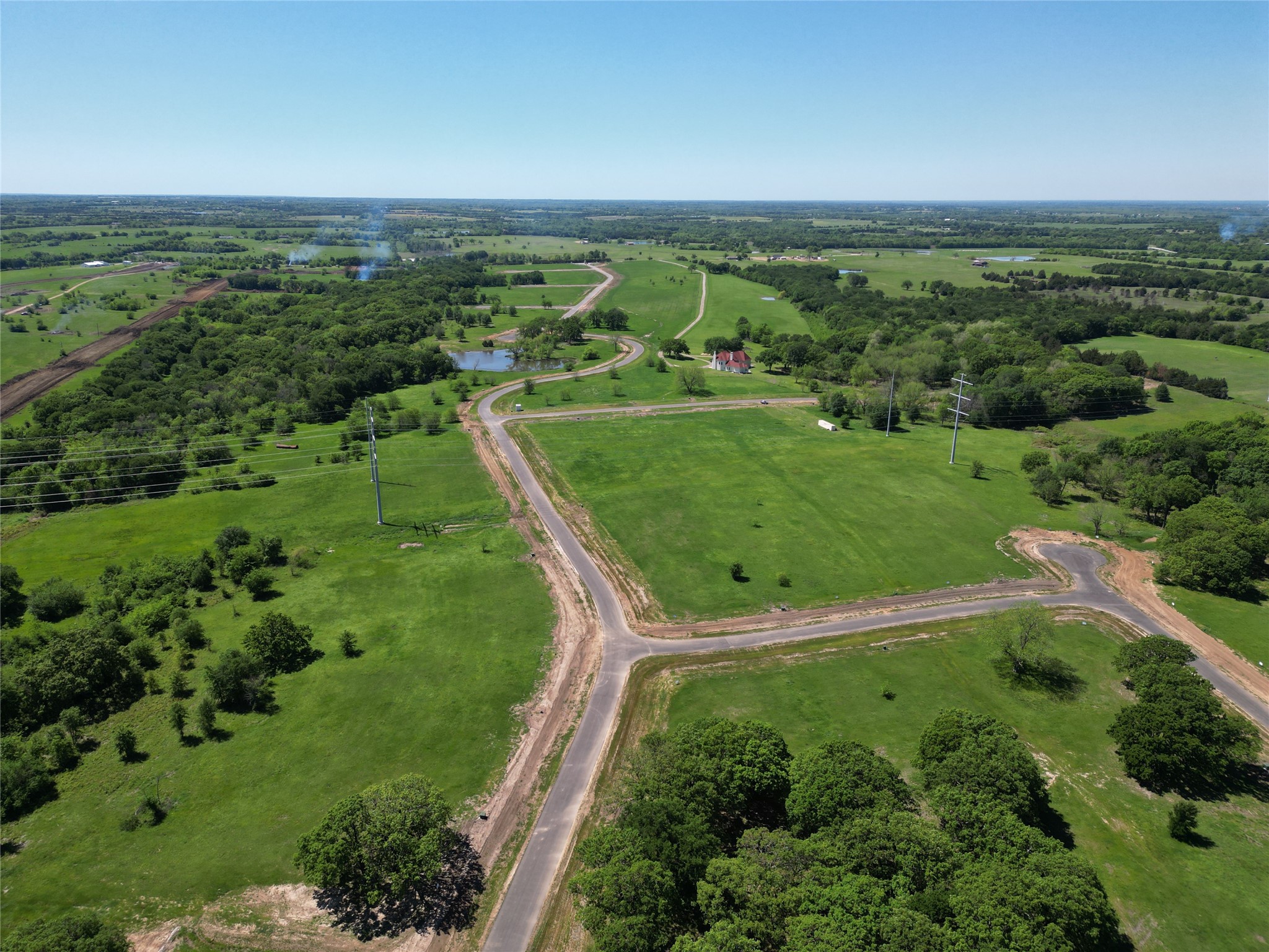 Aerial view of green rural acreage with winding new roads, scattered trees, a pond, and a distant red-roofed house.