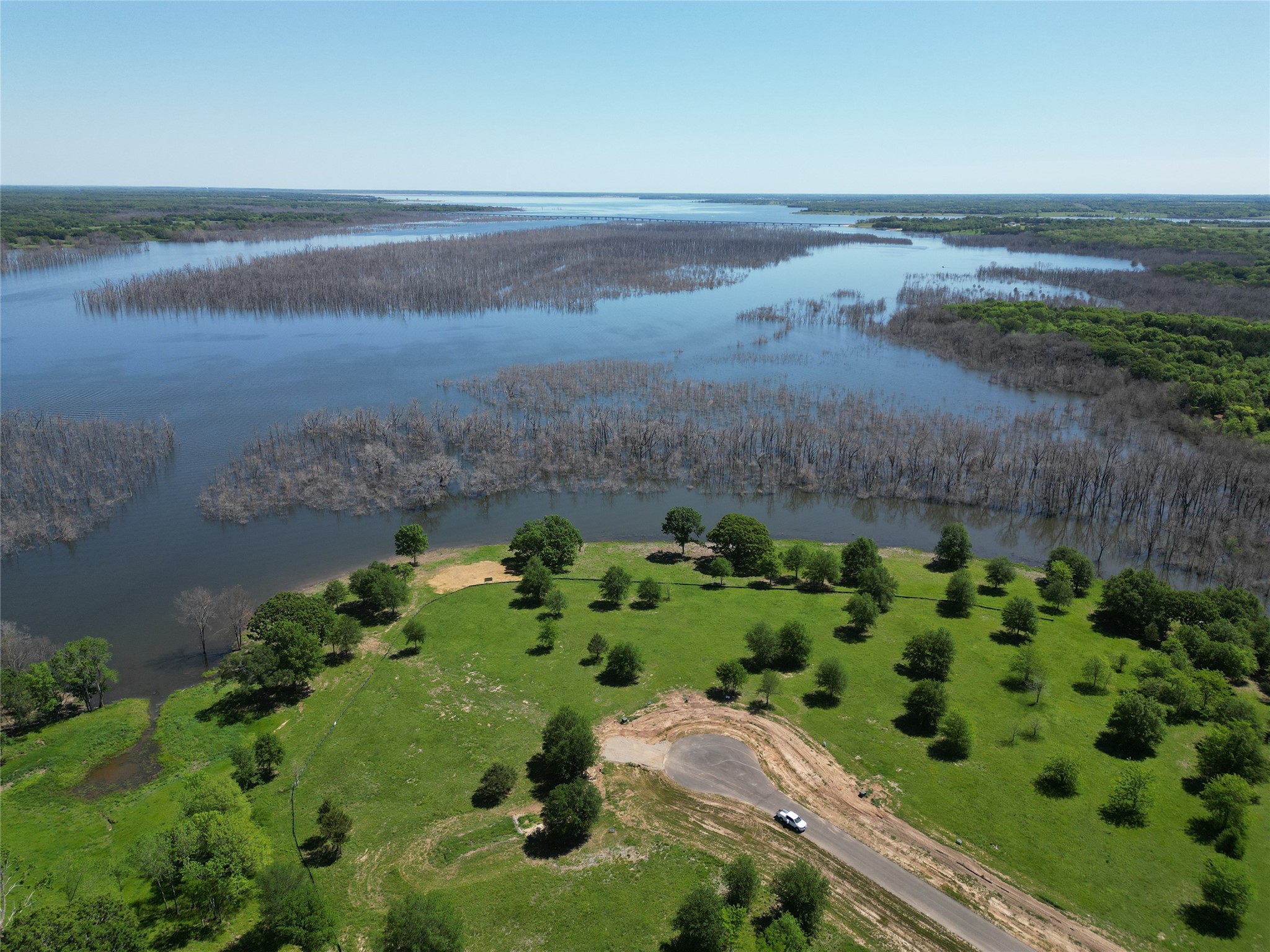 Grassy waterfront property with a cleared circular drive beside a broad, tree-lined body of water.
