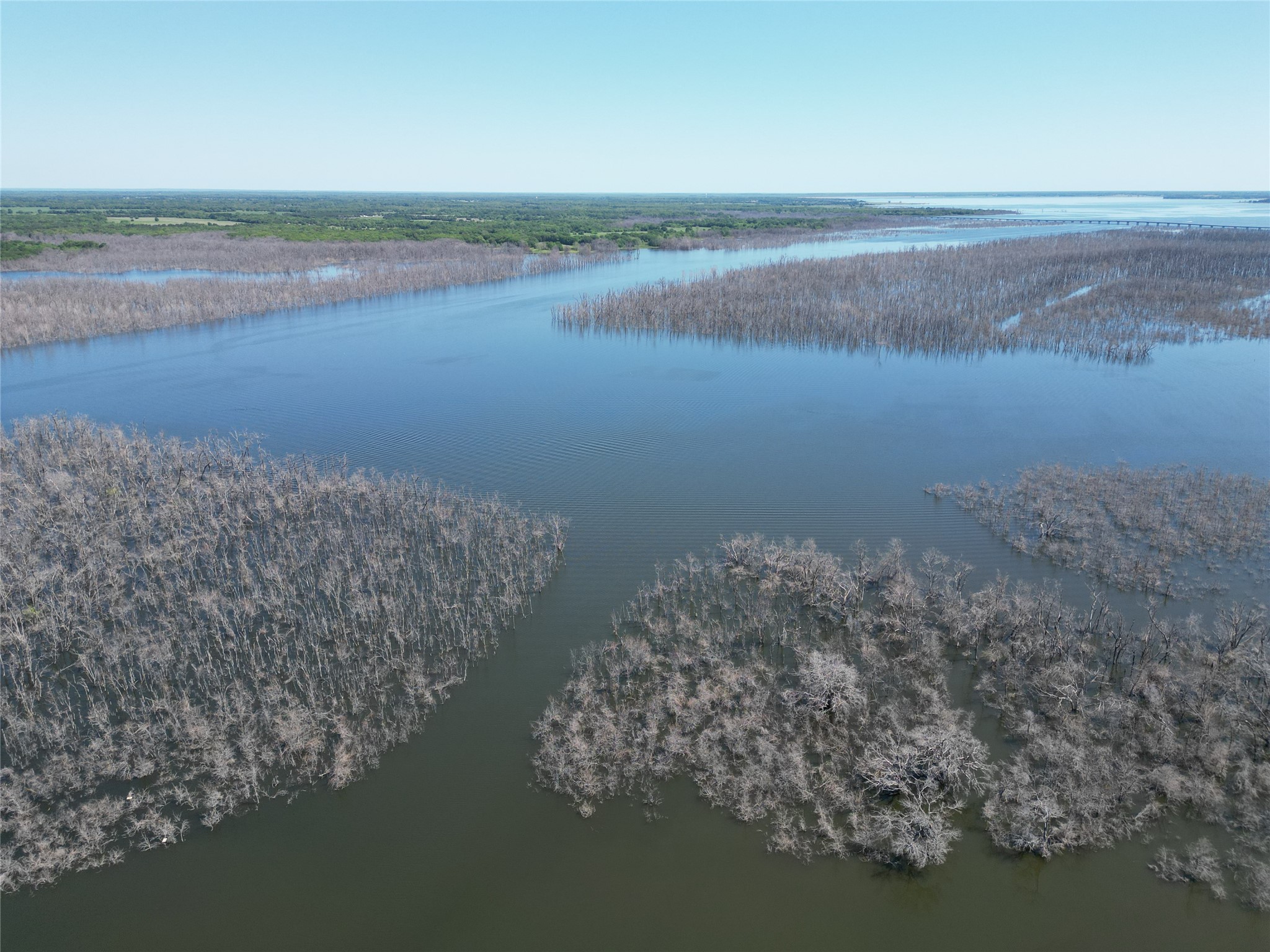 Aerial view over broad water, with wooded wetlands and a distant bridge.