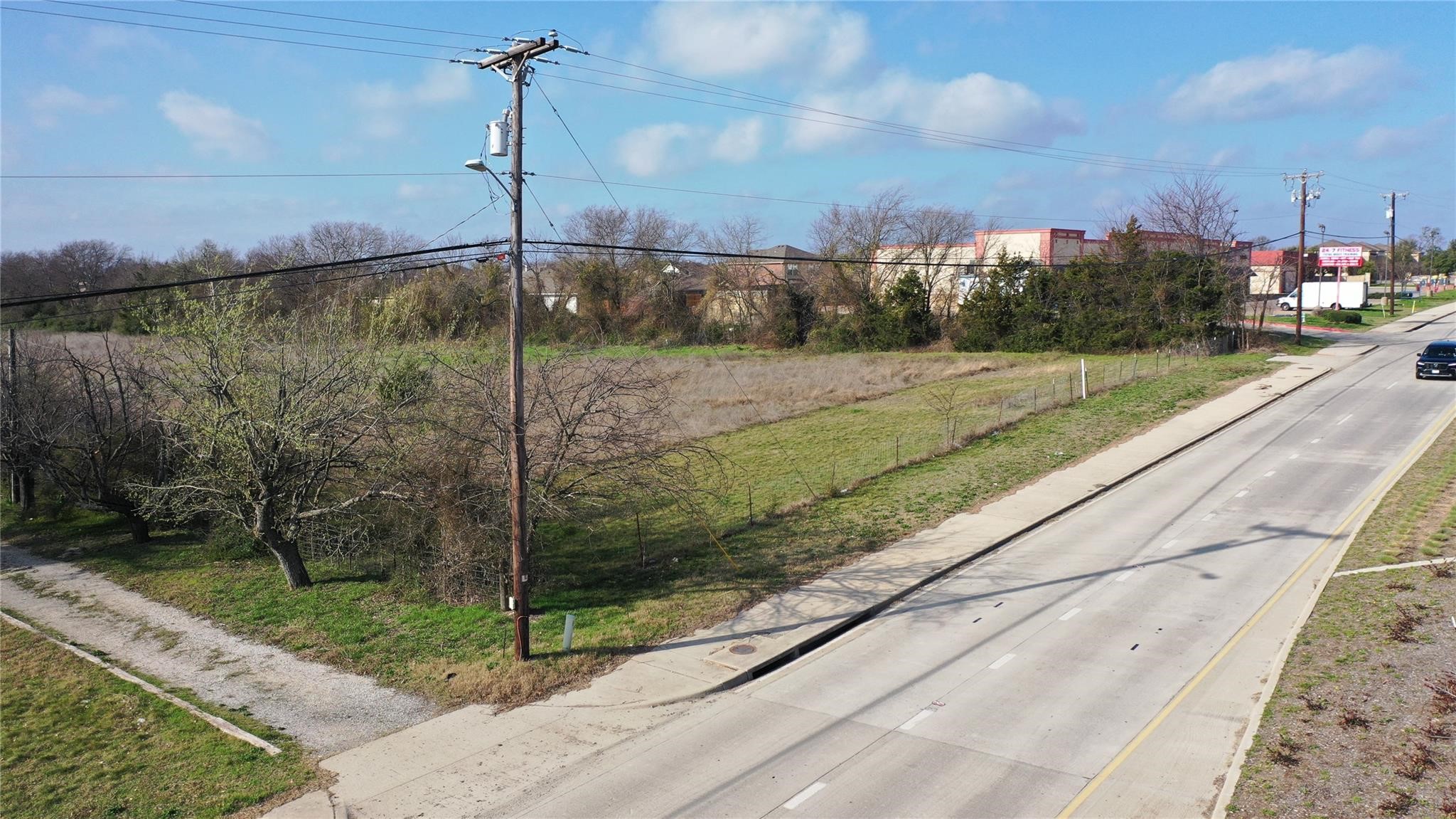 Vacant grassy lot beside a paved road, with scattered leafless trees and commercial buildings nearby.