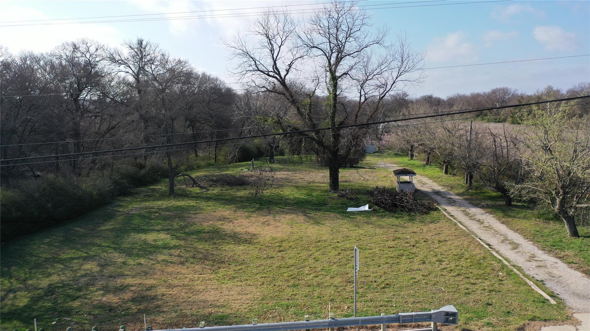 Grassy property lot with a gravel driveway, leafless trees, and a small roofed structure.