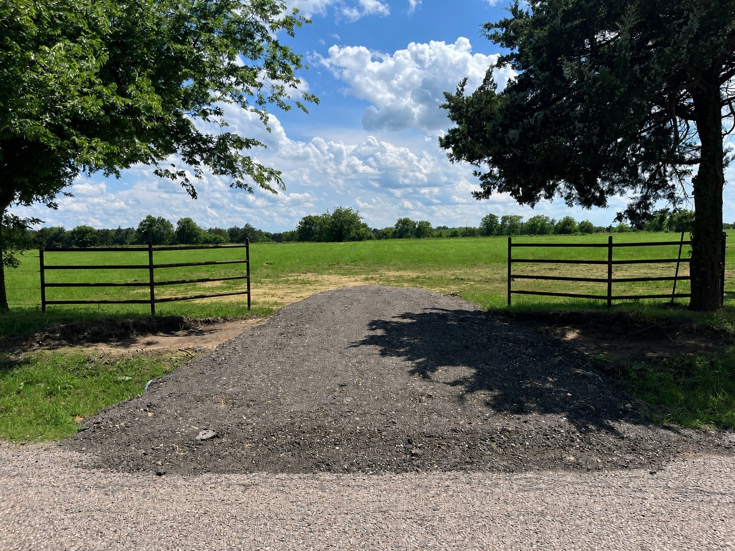 Gravel driveway entrance between metal gates opening onto a green pasture, framed by mature trees.