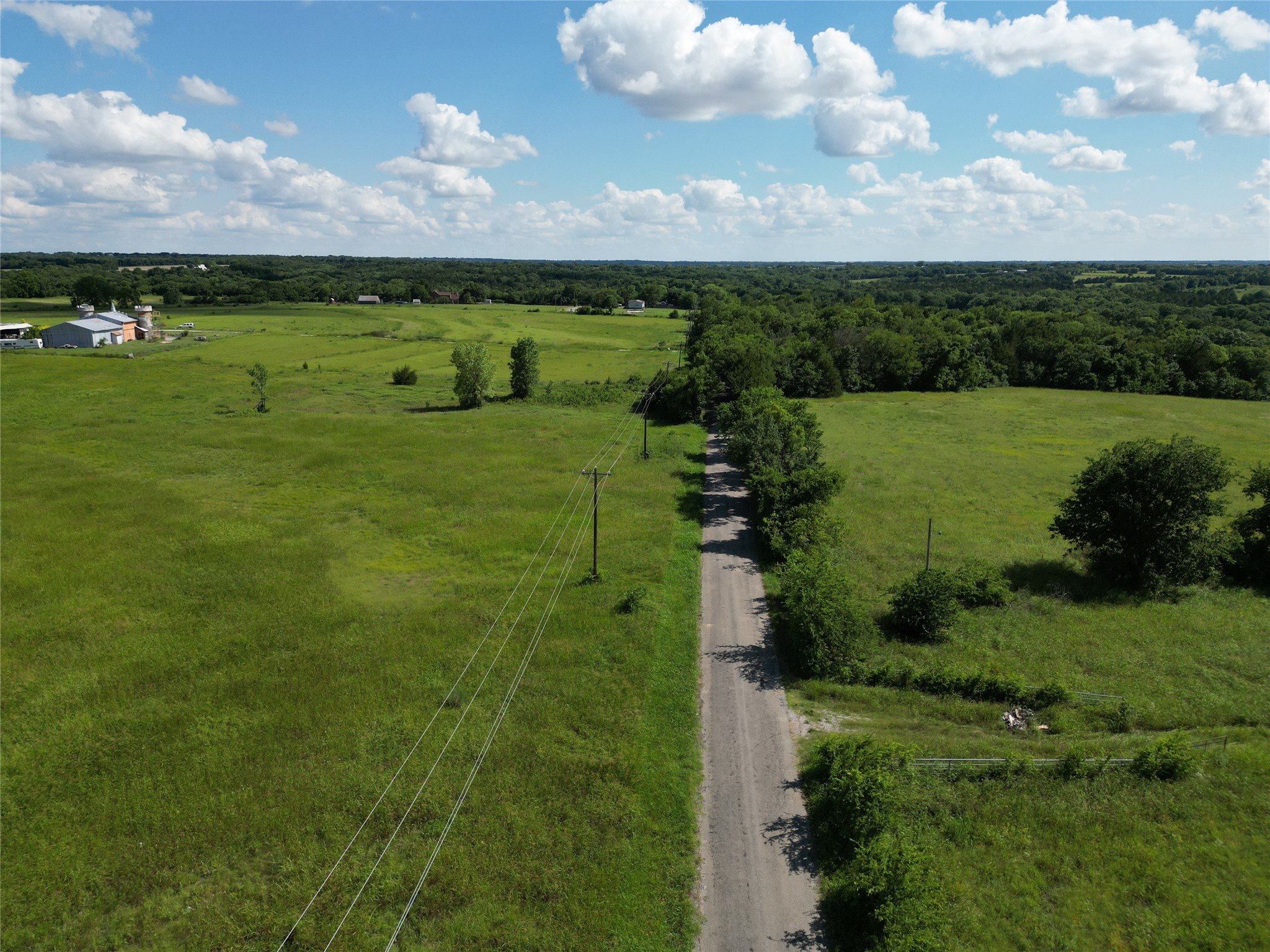 Aerial view of green acreage with a gravel road, utility lines, and a distant barn.