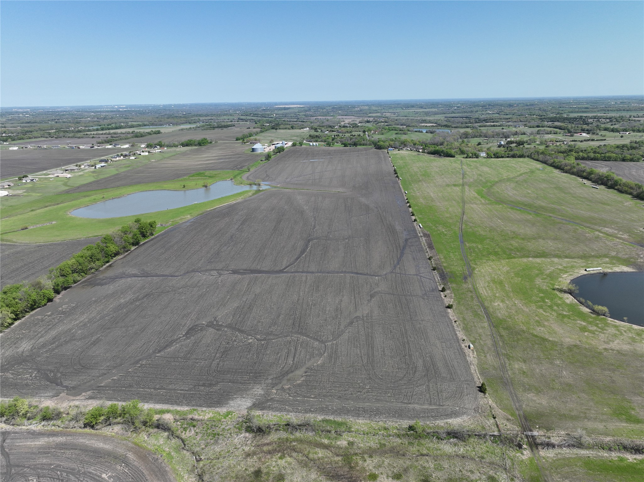 Aerial view of plowed farmland bordered by green pasture, with ponds and scattered farm buildings.