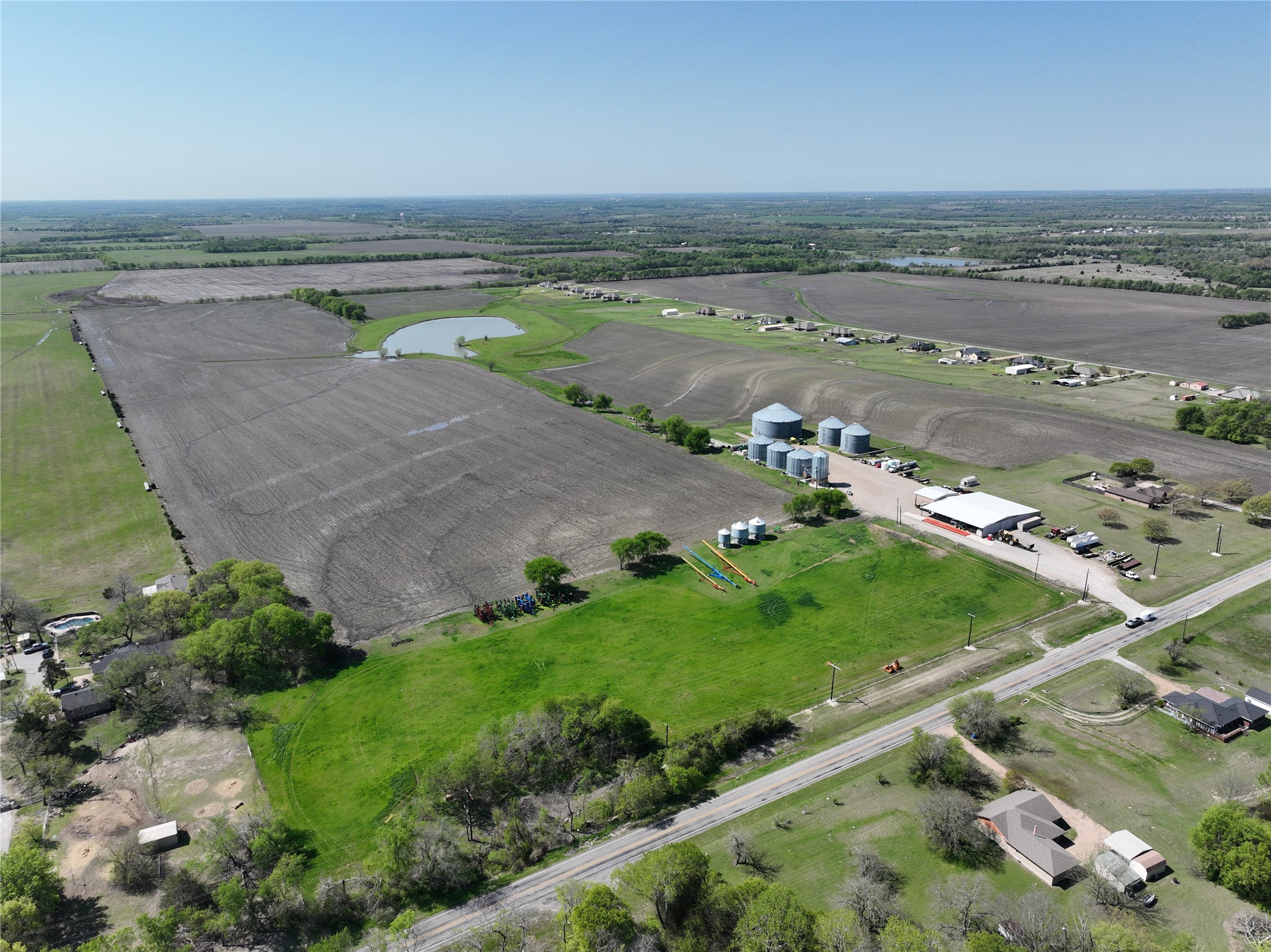 Aerial view of expansive farmland with cultivated fields, grain silos, pond, and roadside buildings.