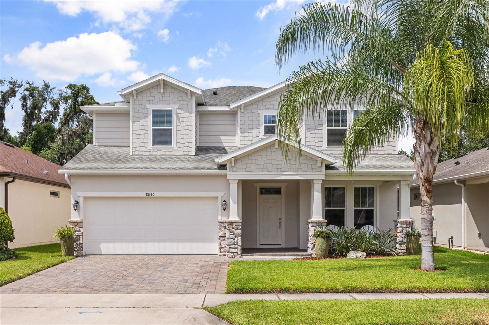 Two-story light-colored home with a wide garage, stone accents, and a palm tree by the driveway.