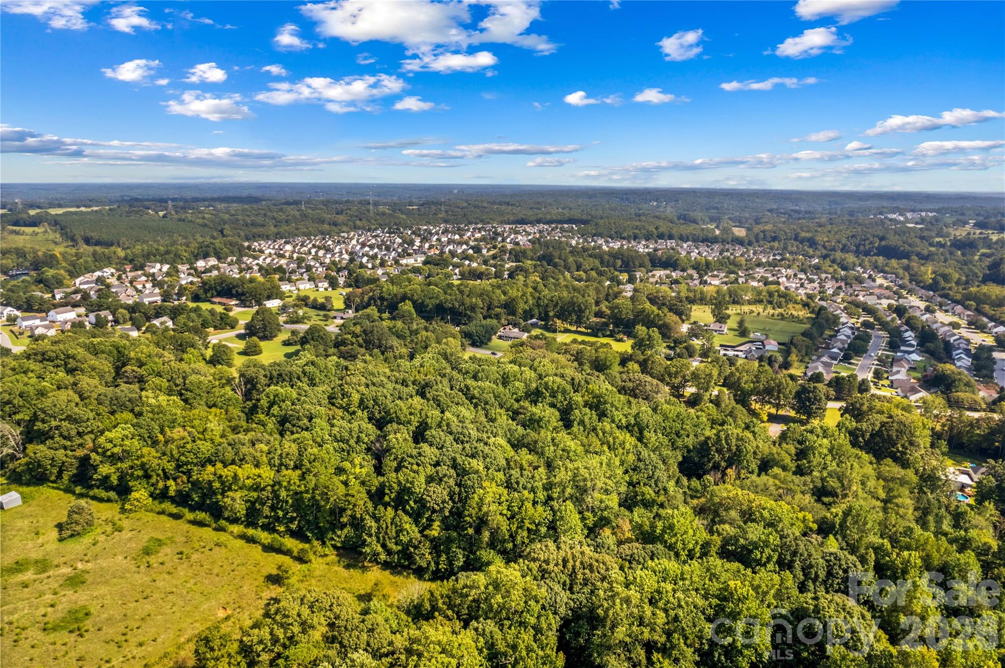 Aerial view of a wooded property beside open grass, with suburban neighborhoods in the distance.