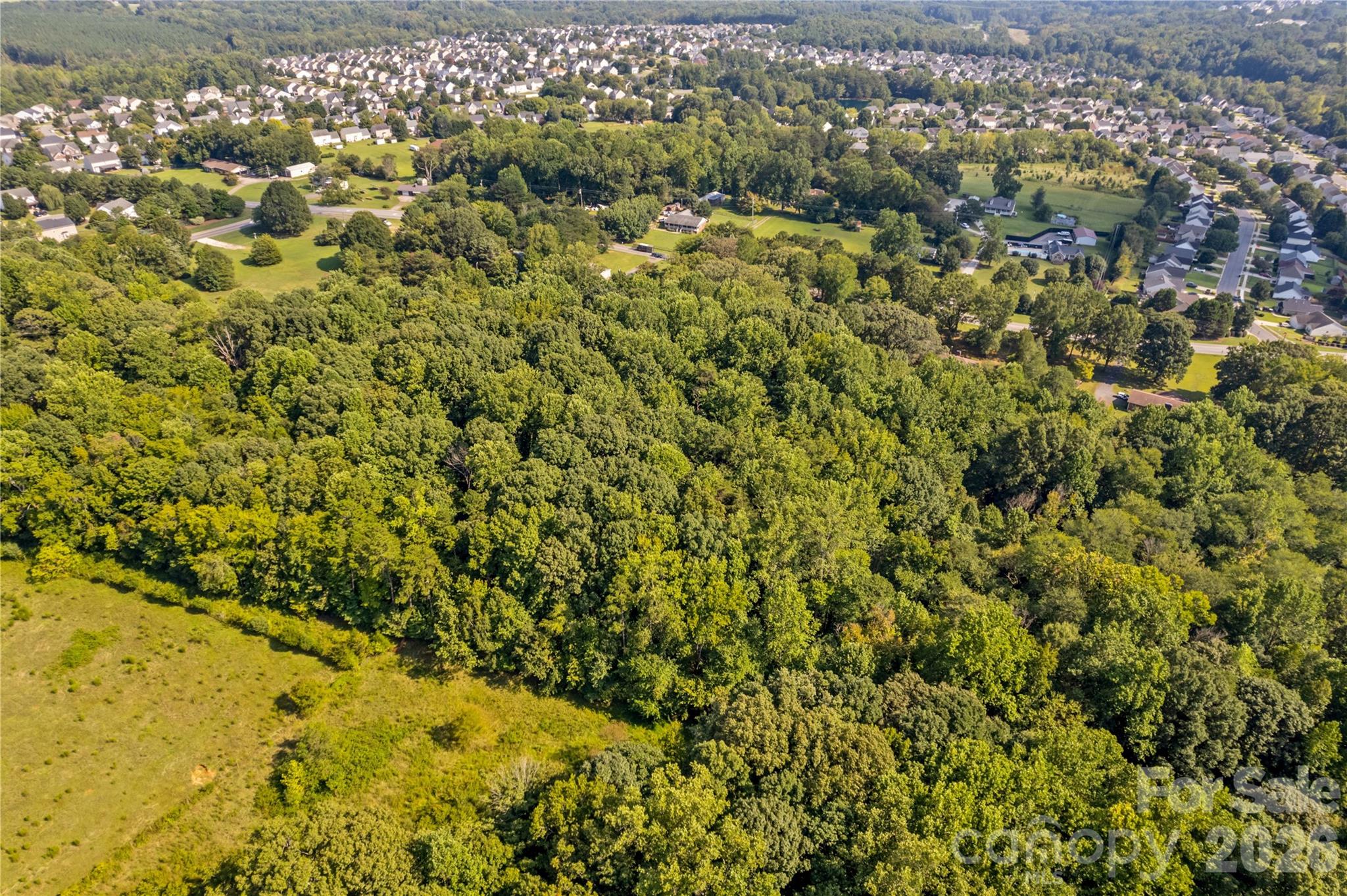 Aerial view of wooded acreage beside a grassy clearing and nearby residential neighborhoods.