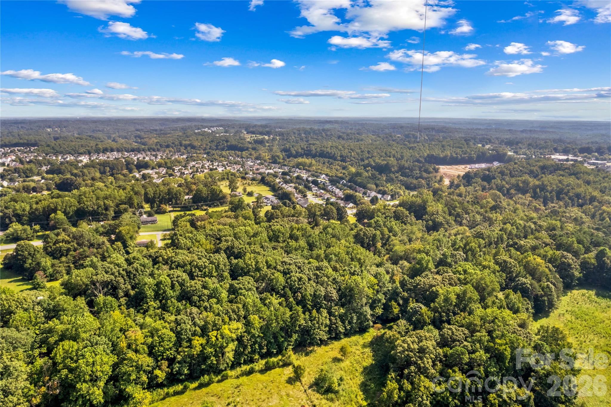 Aerial view of wooded land with a grassy clearing and neighborhood in the distance.