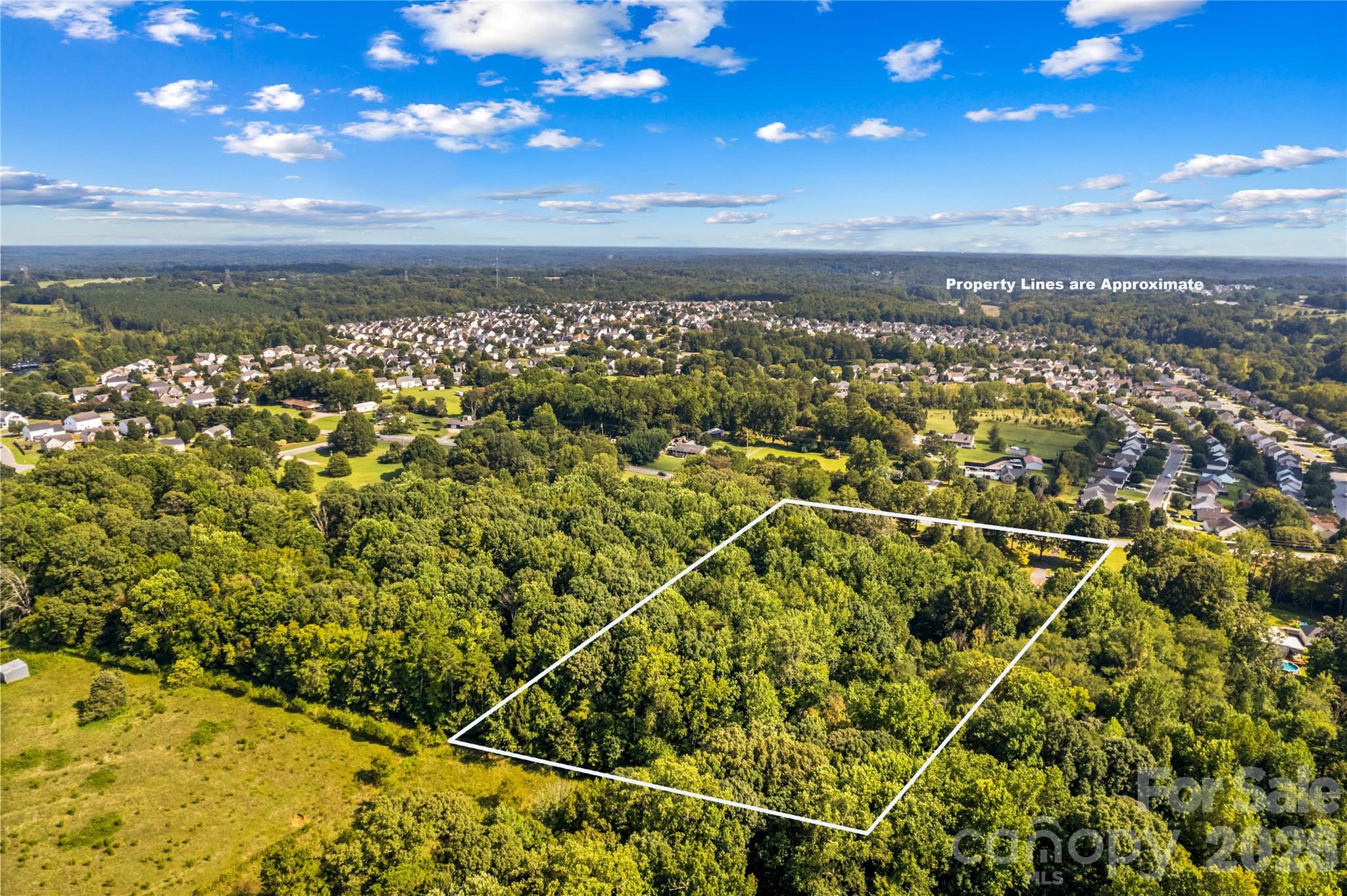 Aerial view of a wooded property outlined by white boundary lines, with neighborhoods and open grass nearby.