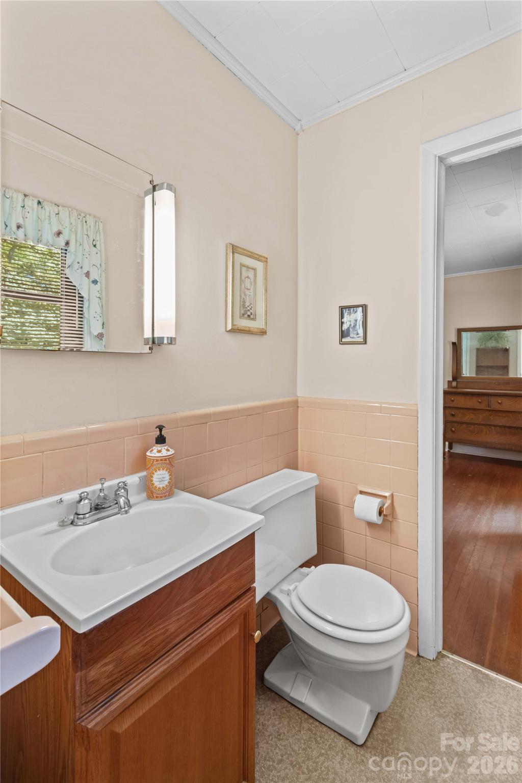 Bathroom with a wood vanity, white fixtures, peach tile, and an open doorway to a hardwood-floored room.