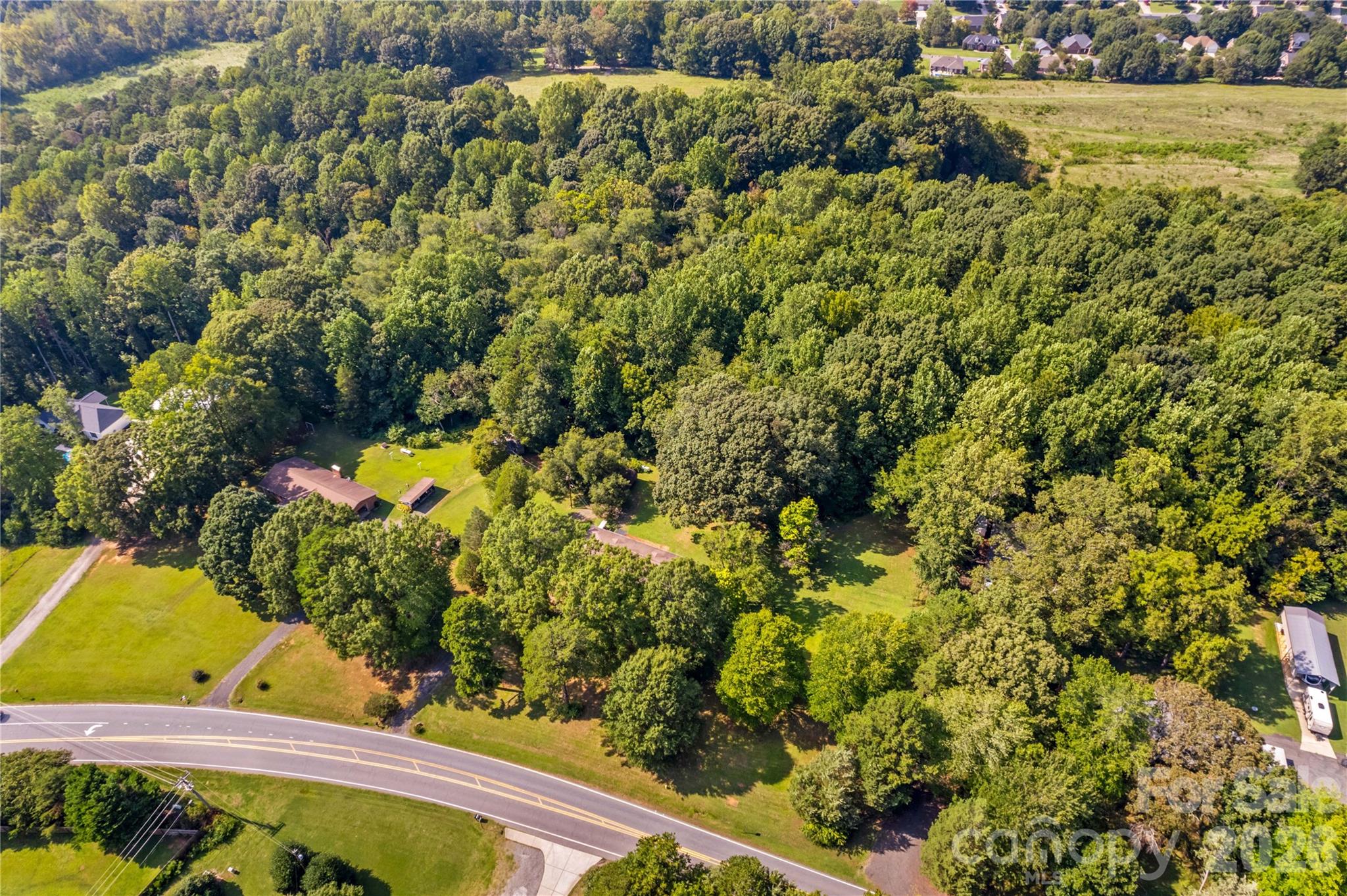 Aerial view of a wooded property with a home, open lawn, and curved road frontage.
