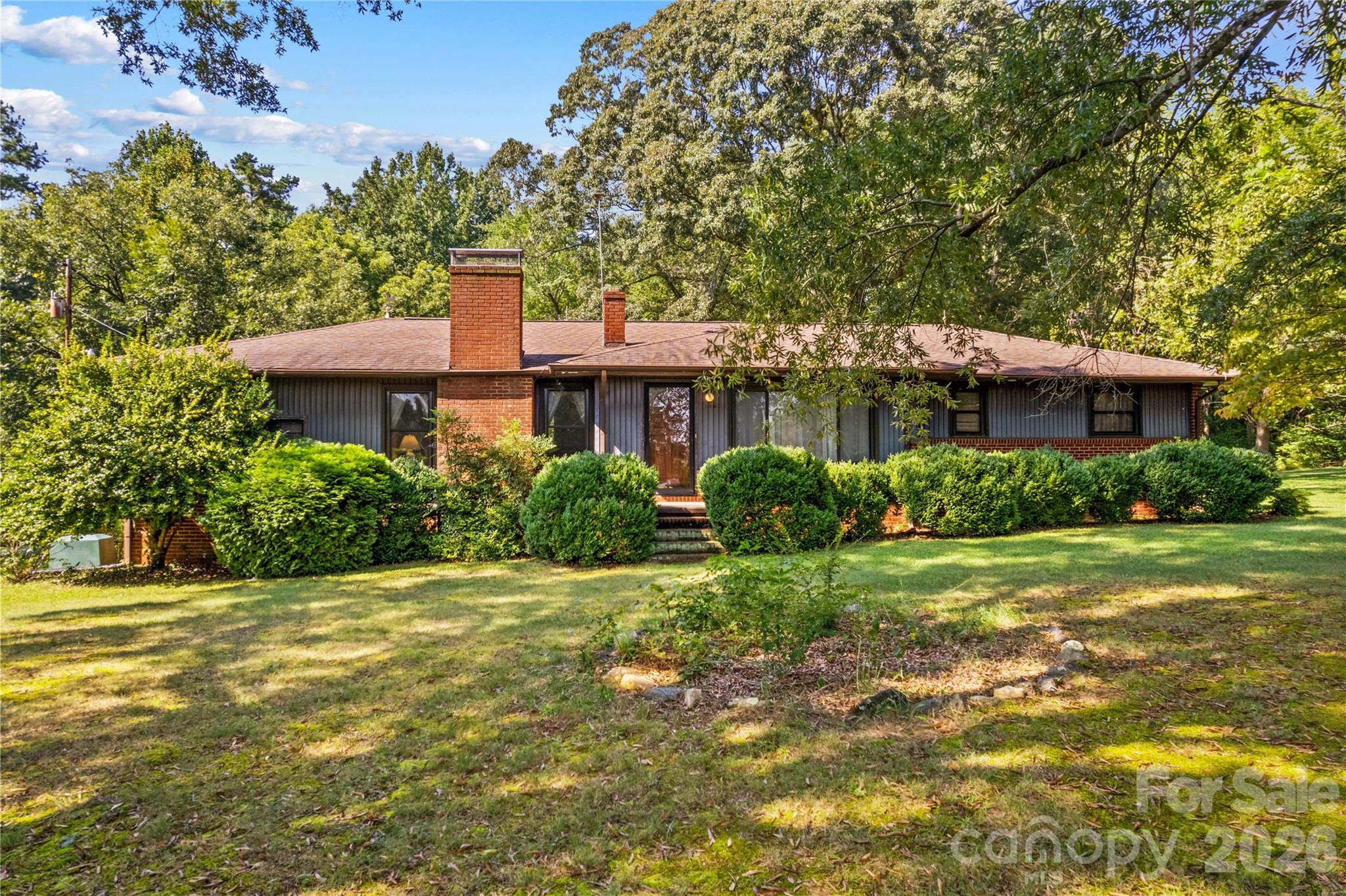 Single-story home with brick chimneys and dark siding, framed by clipped shrubs and mature trees.