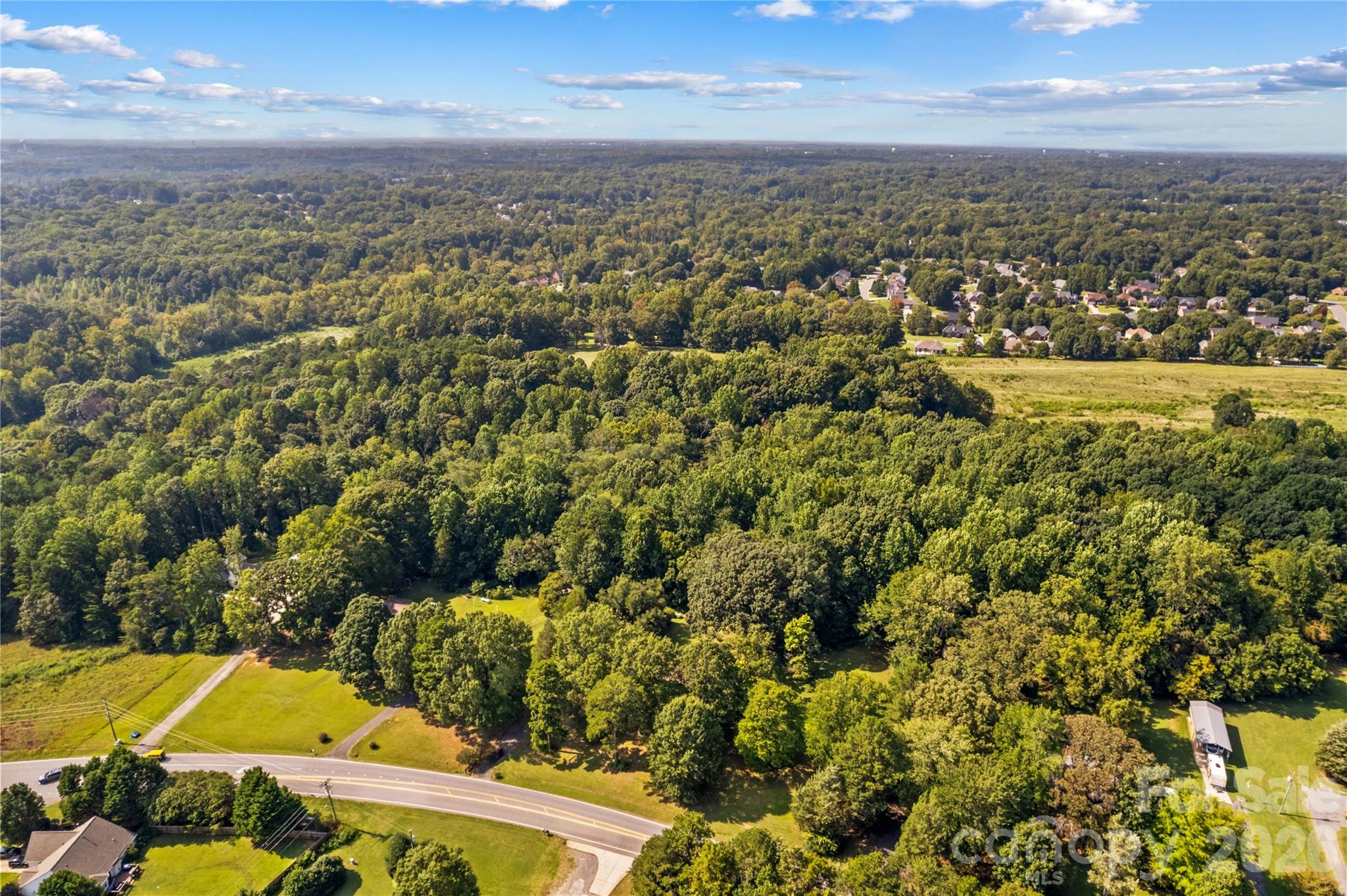 Aerial view of wooded land with open grassy areas, a road, and homes in the distance.