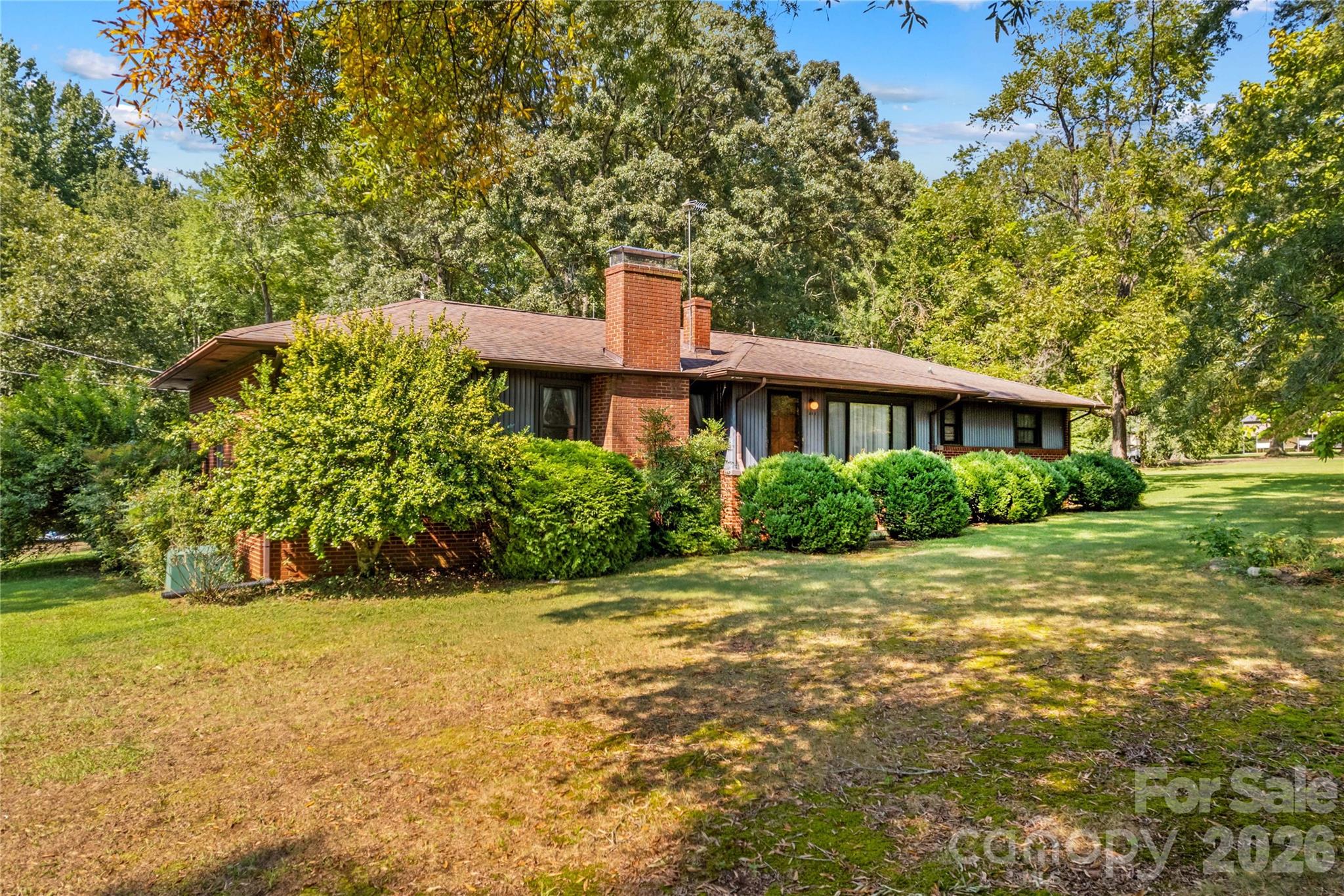 Single-story brick house with a prominent chimney, shrubs, and a broad tree-shaded lawn.