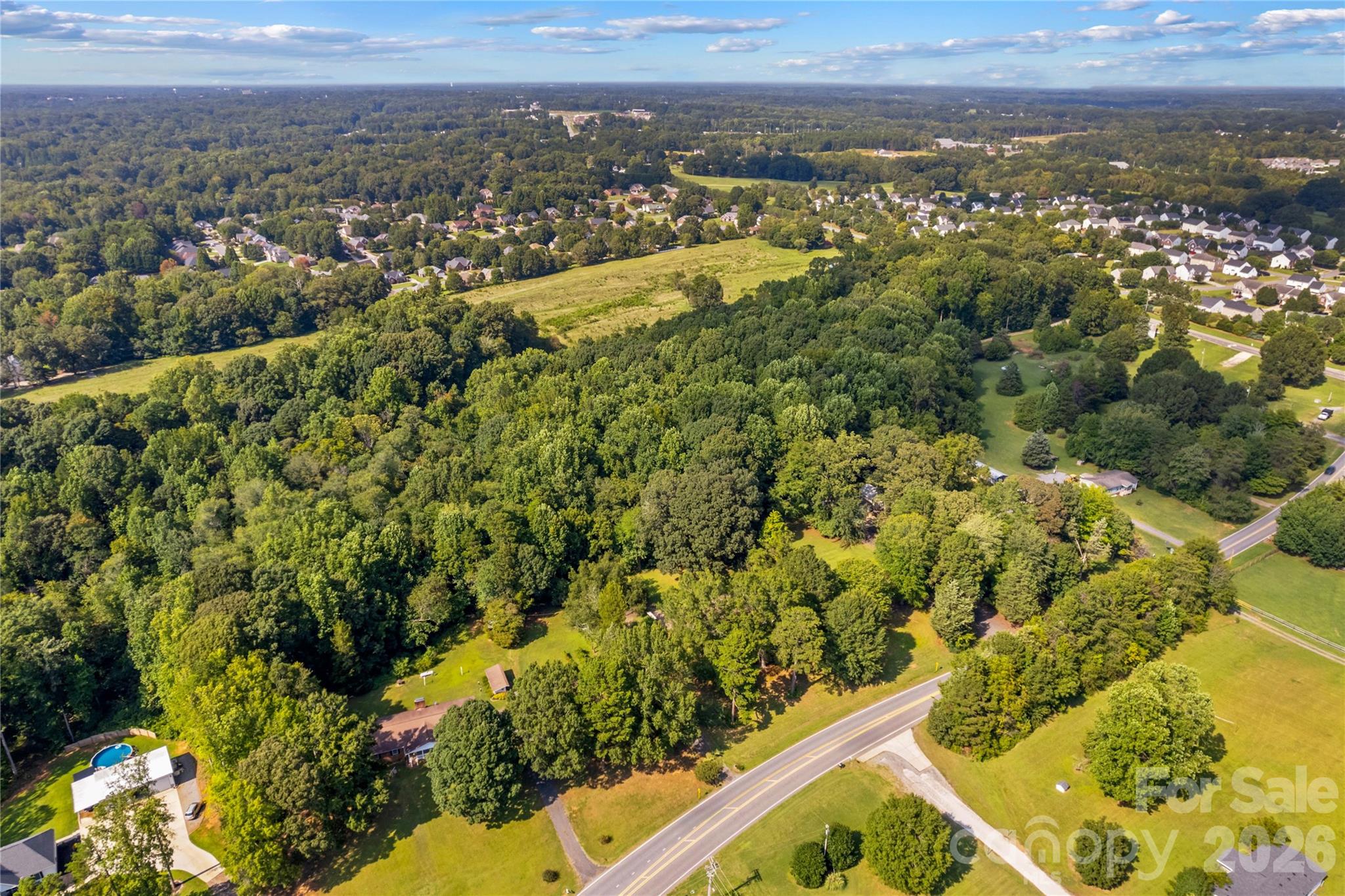 Aerial view of wooded property with a grassy clearing and road frontage beside a suburban neighborhood.