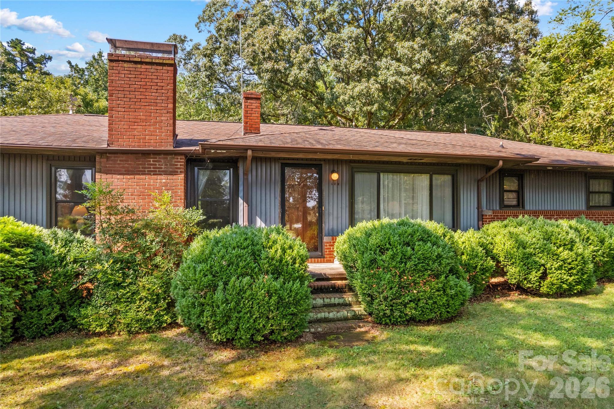 Single-story home with gray siding, red brick chimneys, and rounded shrubs along the front entry.