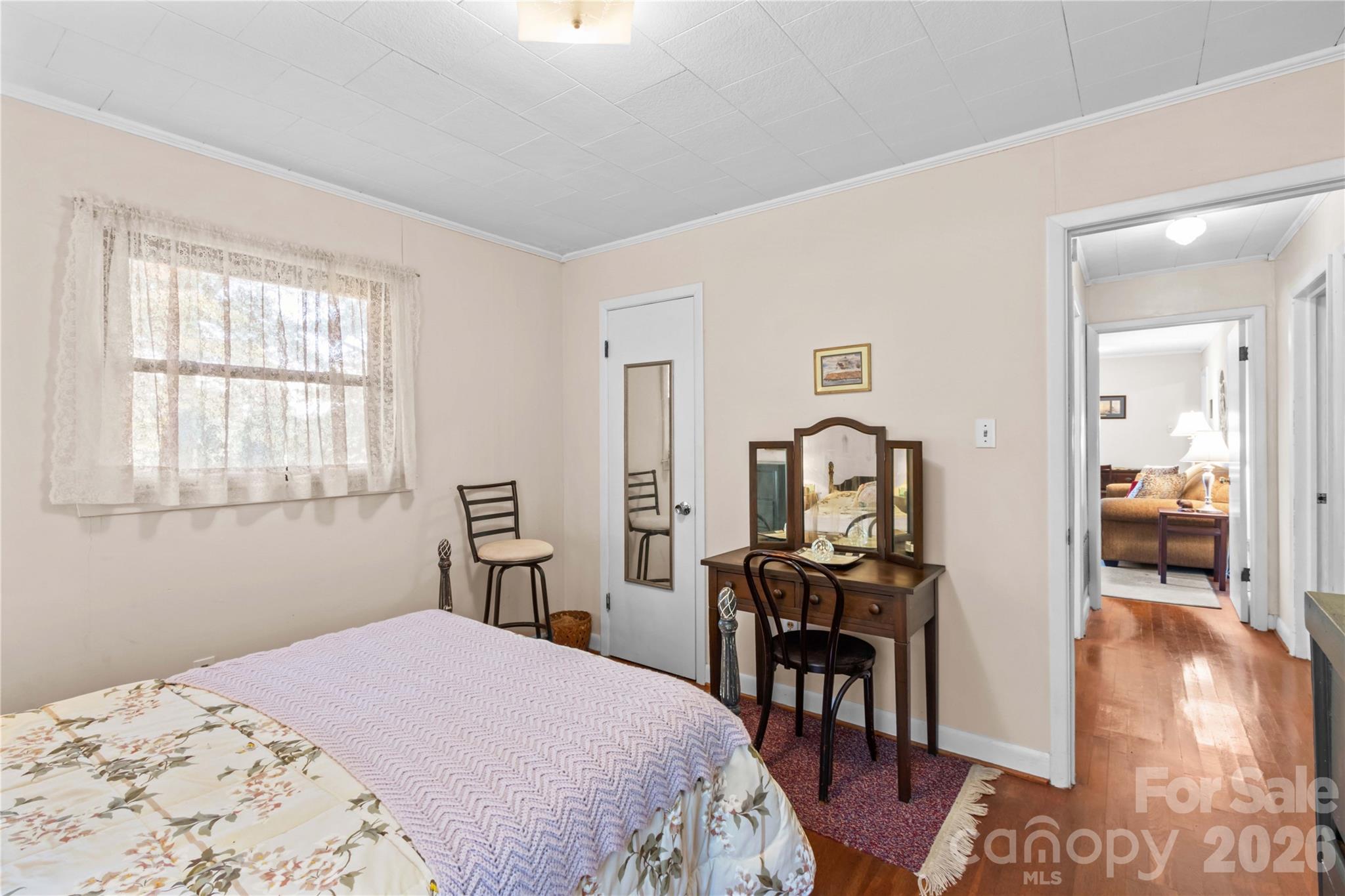 Furnished bedroom with floral bedding, a lace-curtained window, and a wooden vanity beside the hallway.