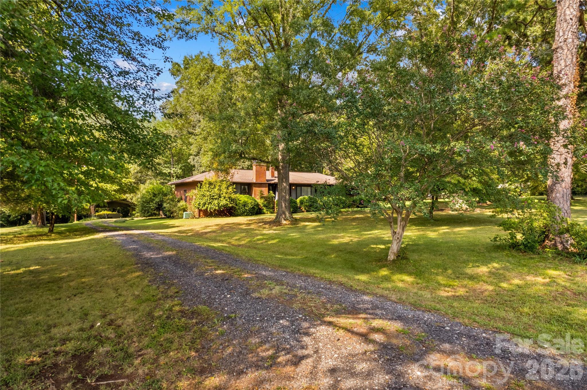 Single-story brick house among mature trees, with a gravel driveway crossing the lawn.