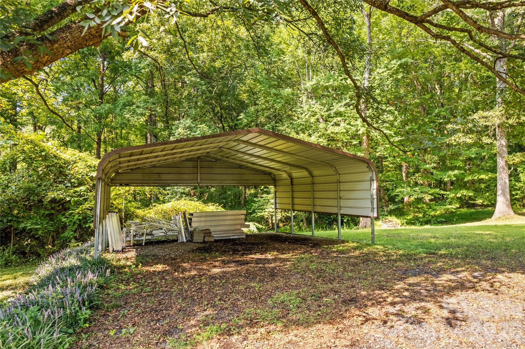 Open metal carport beneath mature trees, with building materials stored underneath.