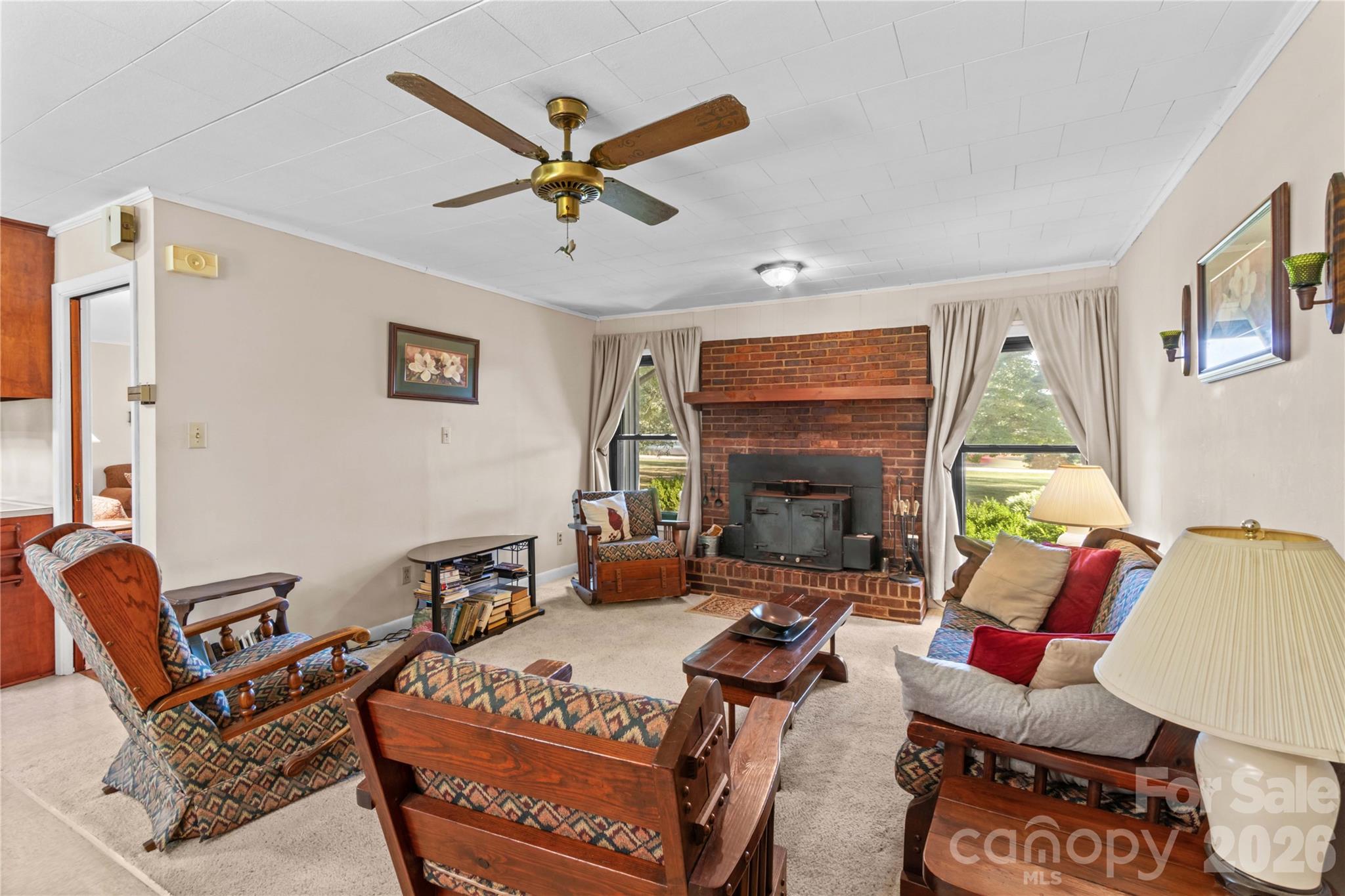 Living room with a brick fireplace, wood-burning stove, patterned wooden chairs, and curtained windows.