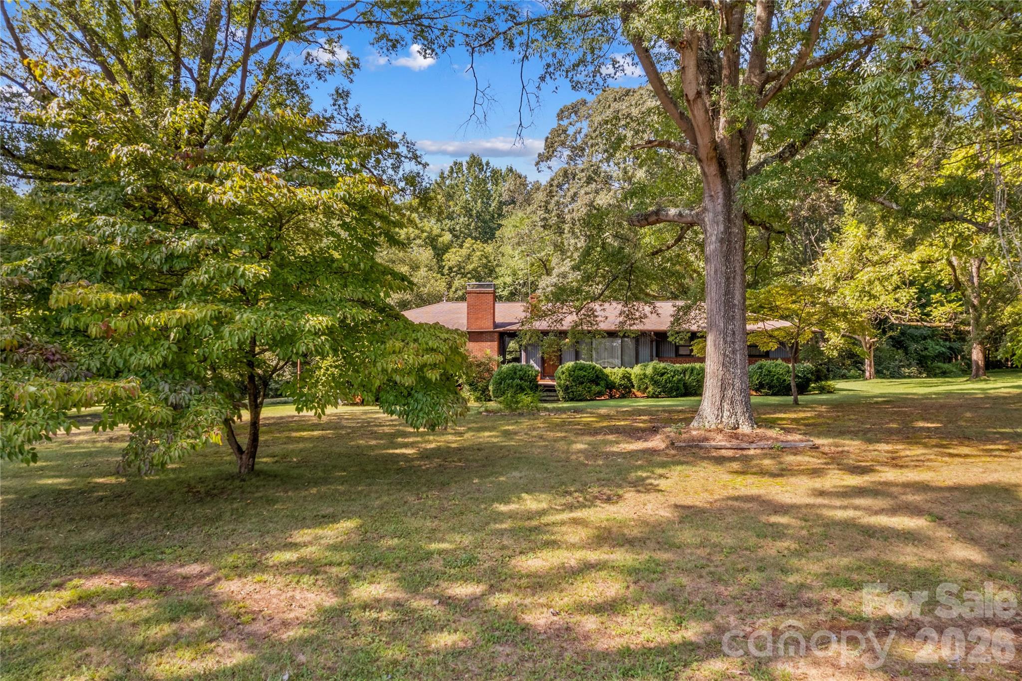 Single-story brick home with a chimney, framed by mature trees across a spacious lawn.