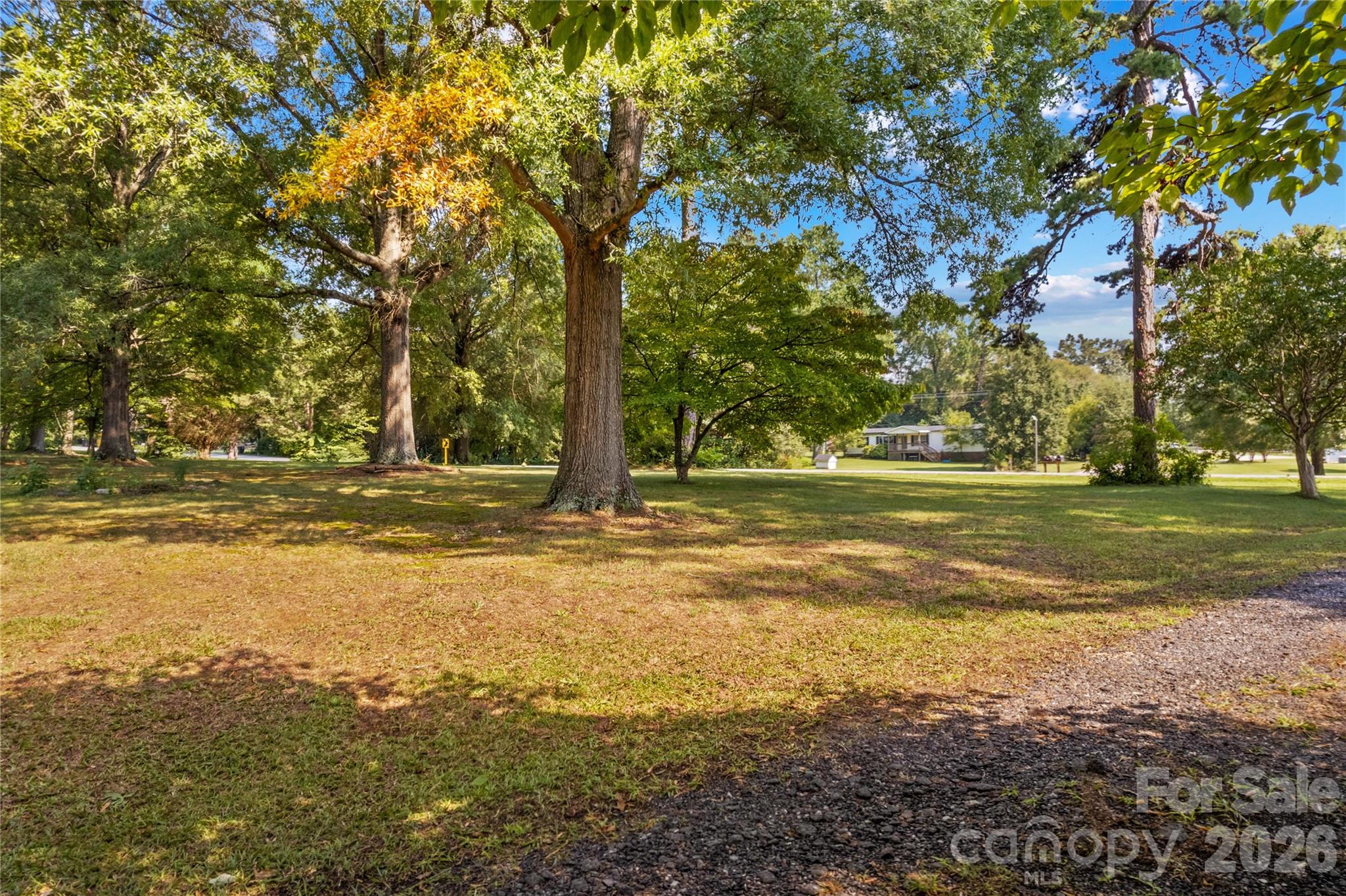 Tree-shaded property lot with a gravel drive and a home across the road.