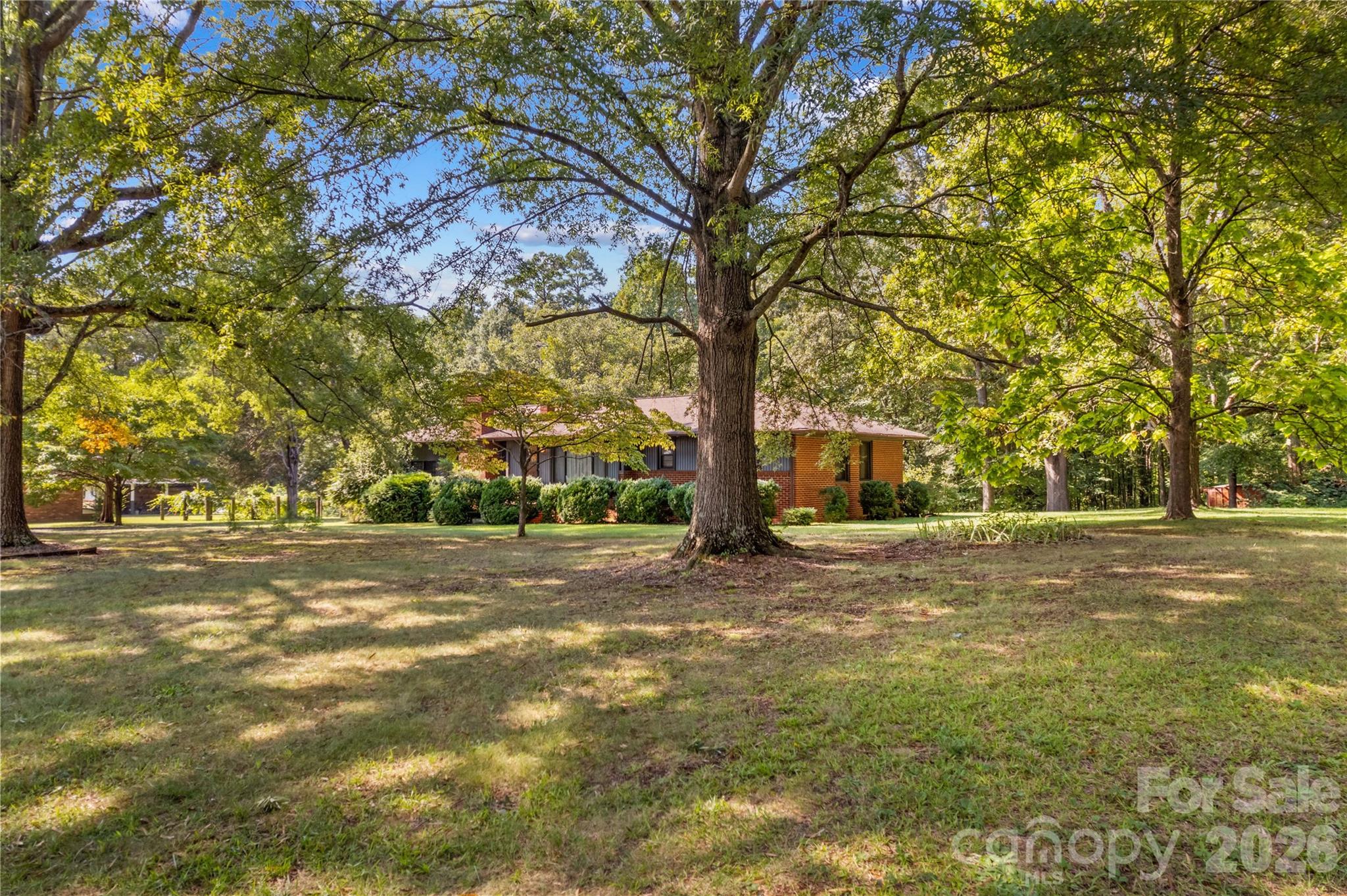 Single-story brick house partly screened by mature trees across a broad, shaded lawn.