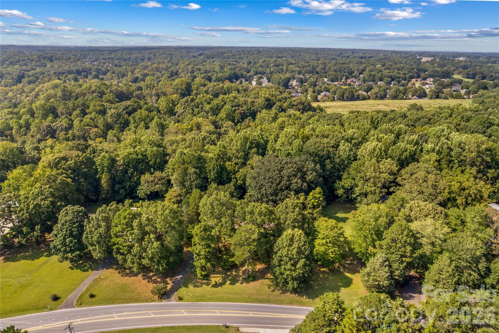 Aerial view of wooded property beside a road, with lawns and a neighborhood beyond.