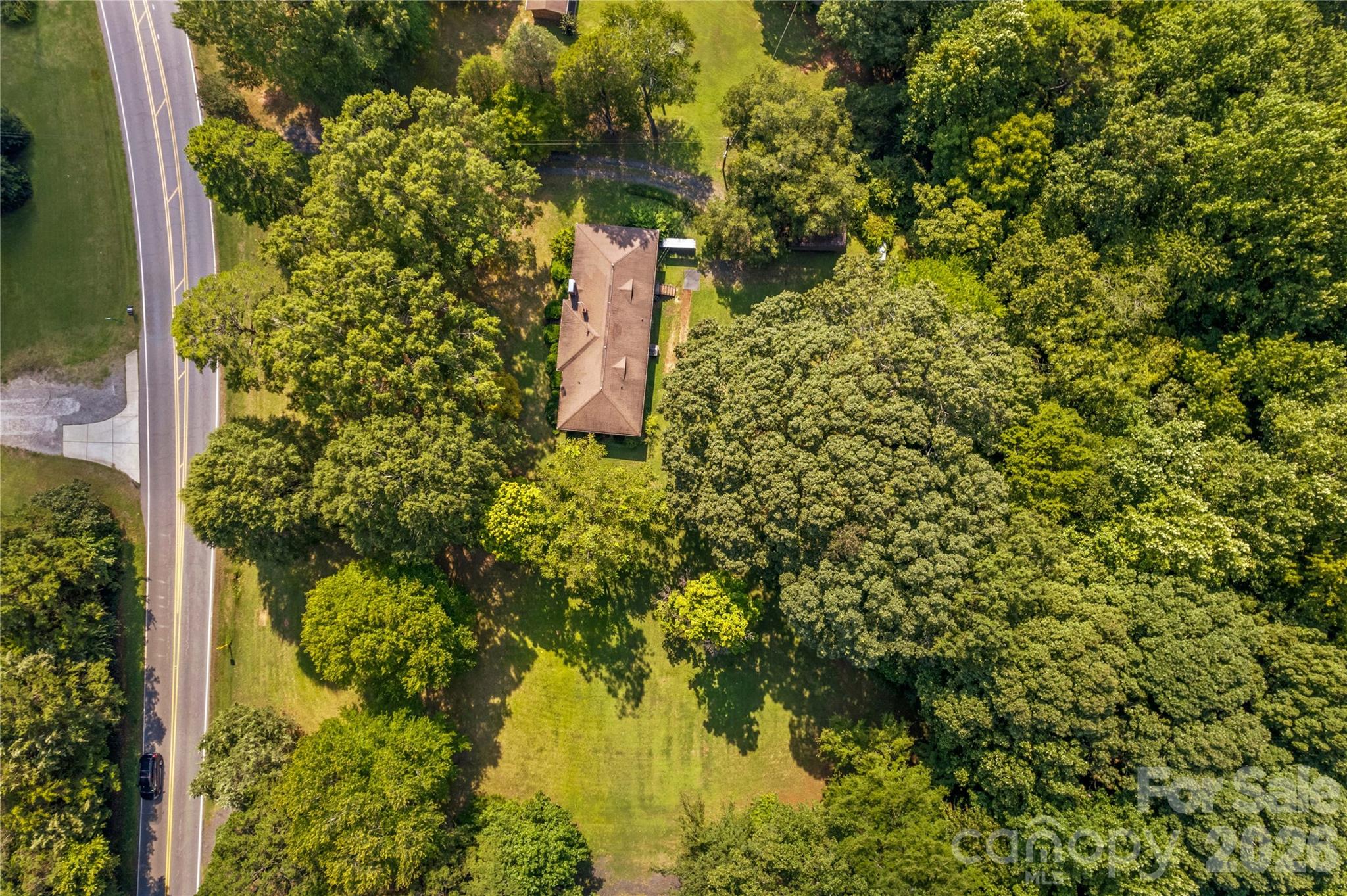 Aerial view of a house on a wooded lot beside a two-lane road, with a broad grassy lawn.