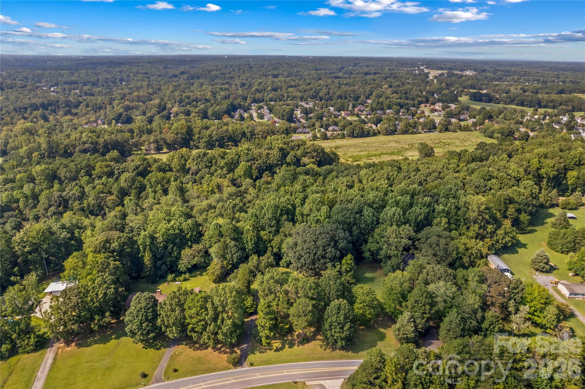 Aerial view of wooded land along a road, with nearby homes and open fields.