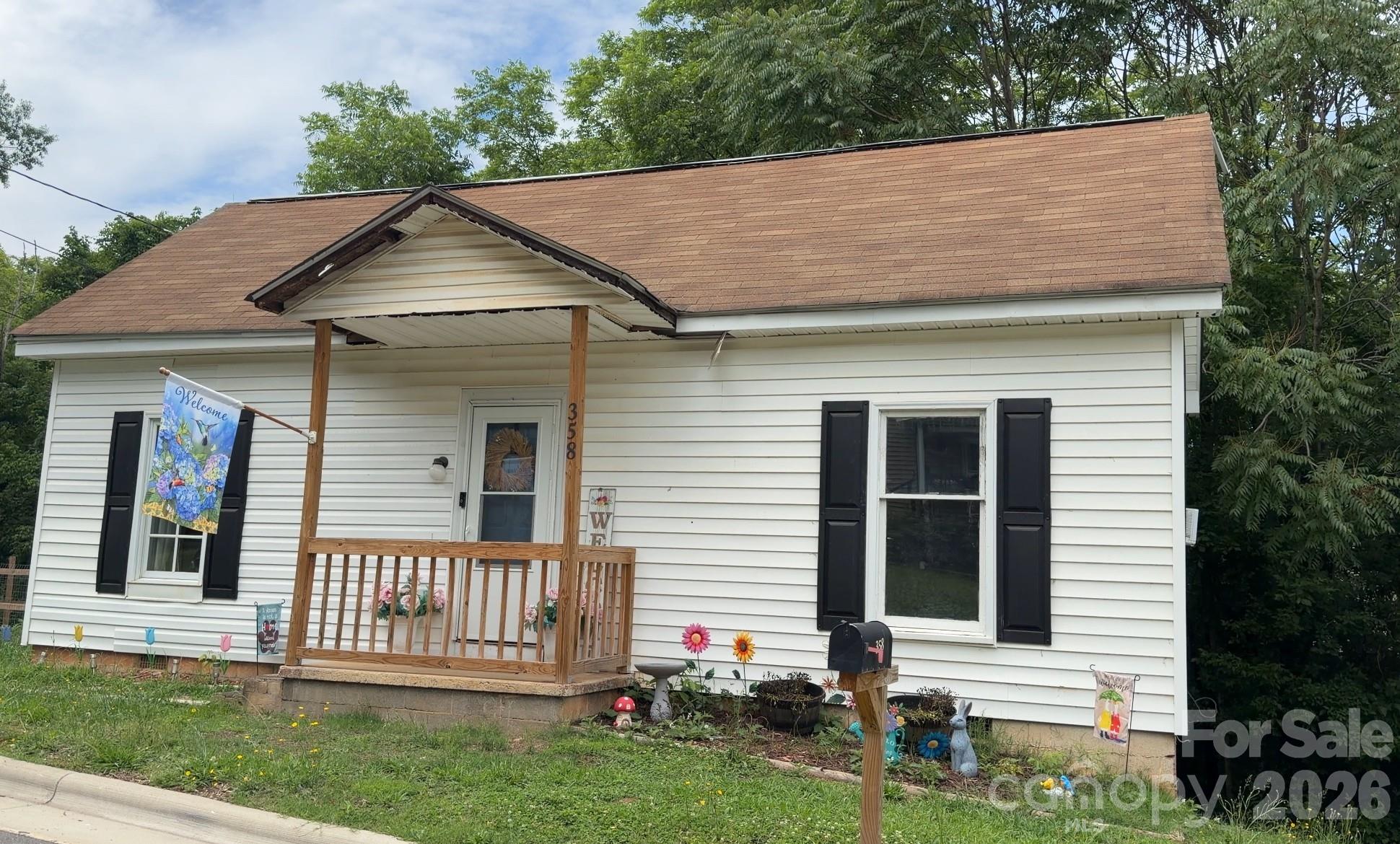 White single-story house with a covered wooden porch, black shutters, and a flower-decorated front yard.