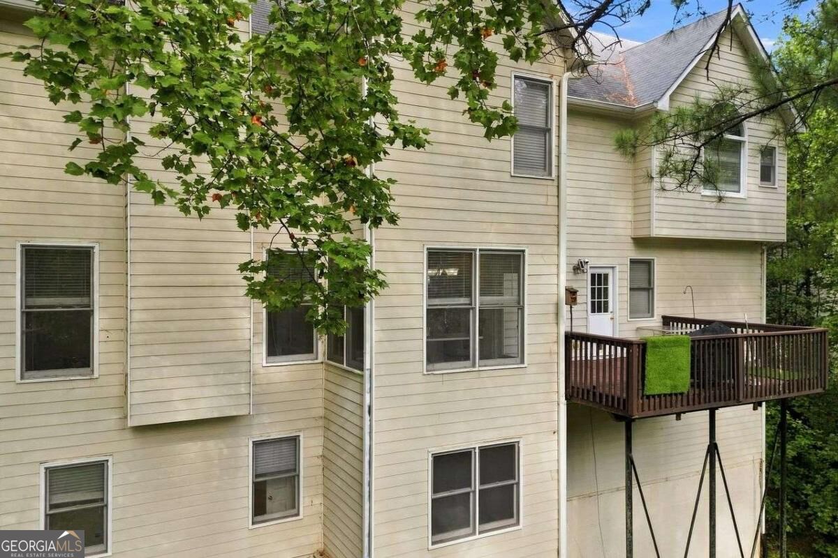 Rear of a beige multi-story house with an elevated wooden deck, partly screened by leafy trees.