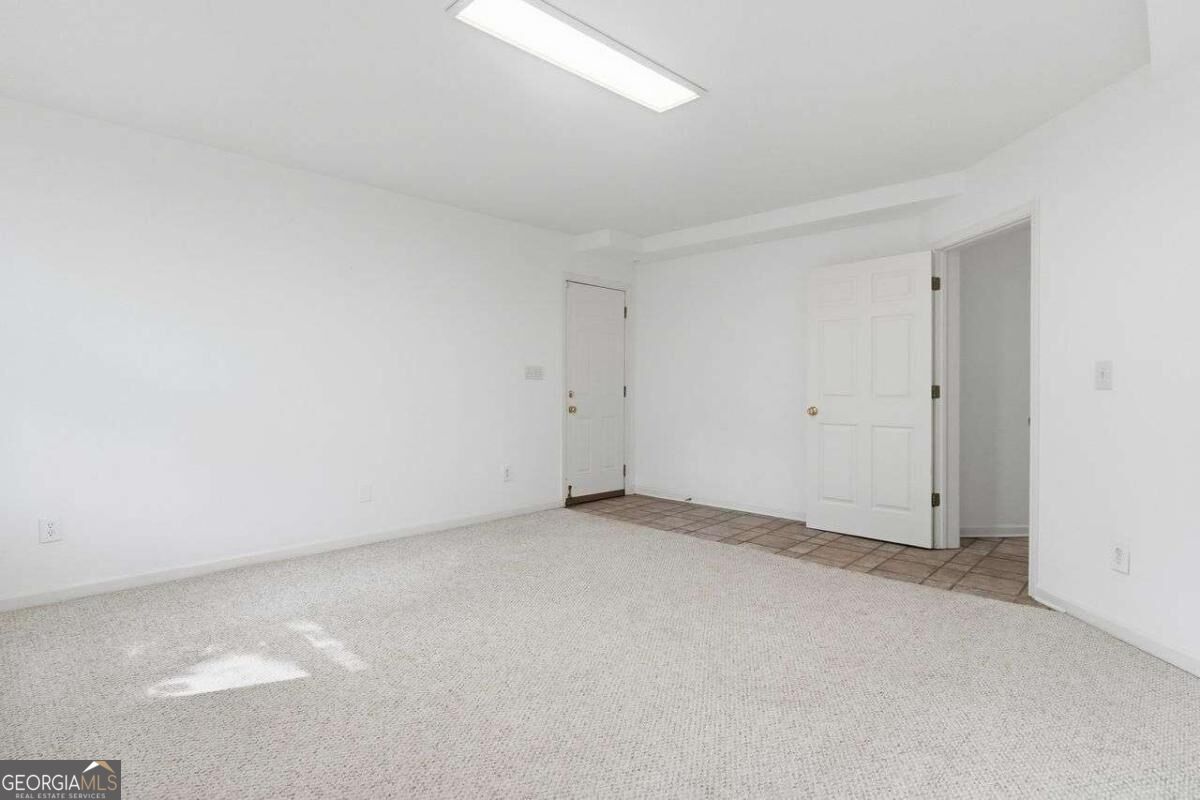 Empty carpeted room with white walls, a ceiling light, and an open doorway beside a tiled area.