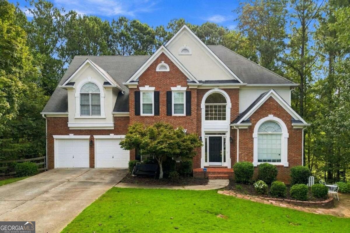 Two-story brick house with a two-car garage, arched windows, and a green lawn surrounded by trees.