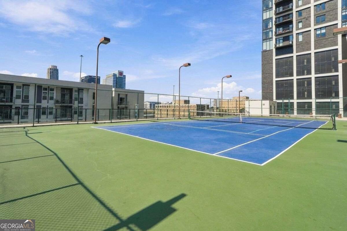 Rooftop tennis court with a blue playing surface, surrounded by urban buildings.