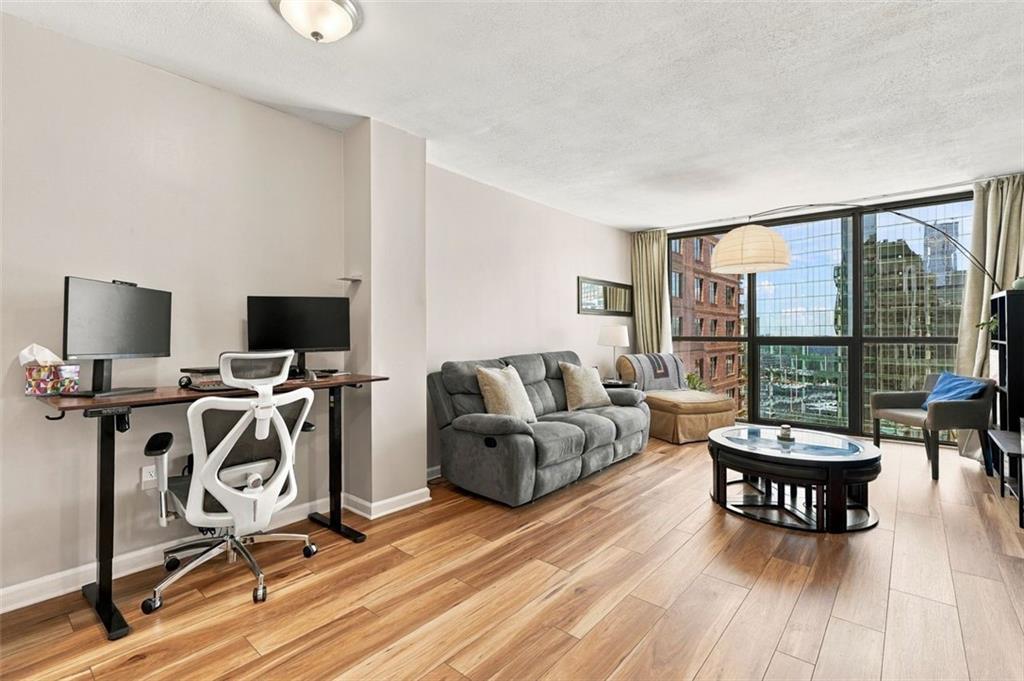 Living room with wood floors, gray sofa, desk, and large windows overlooking city buildings.