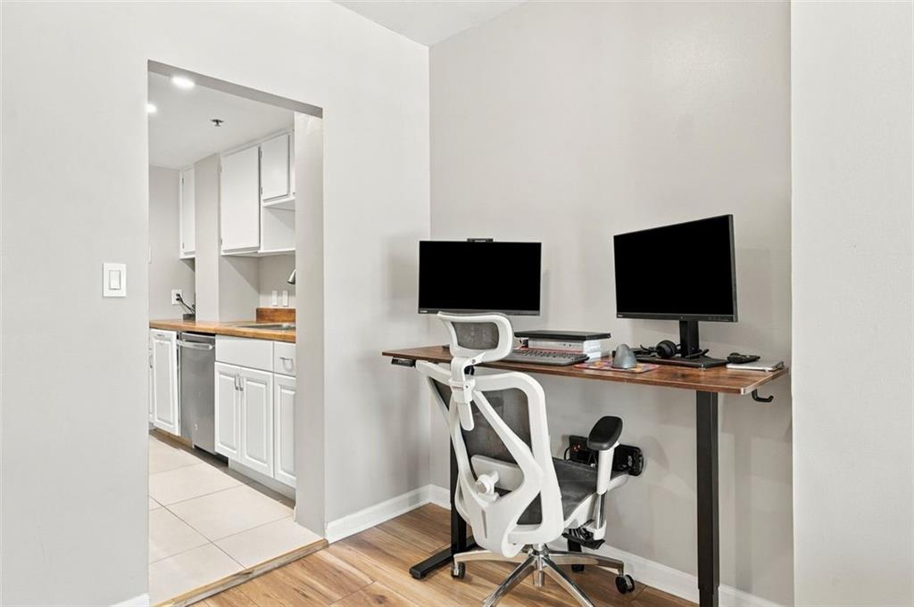 Home office with a two-monitor standing desk and a compact kitchen through the doorway.