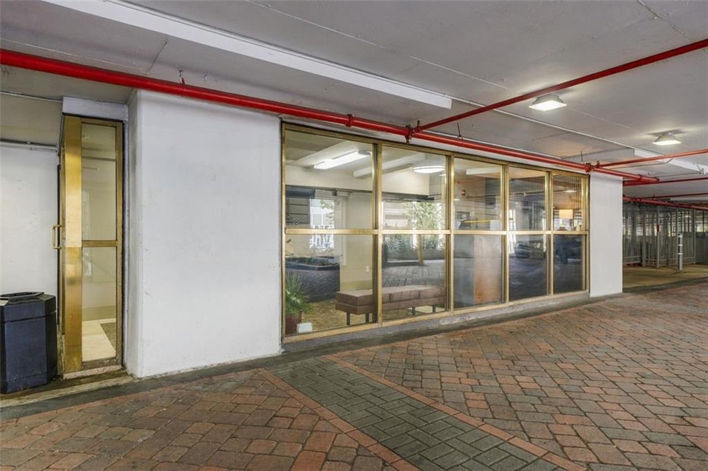 Glass-fronted building facade along a covered walkway with brick paving and red ceiling pipes.