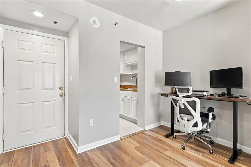 Condo home office with a desk and chair beside the front entry, with the kitchen visible through a doorway.