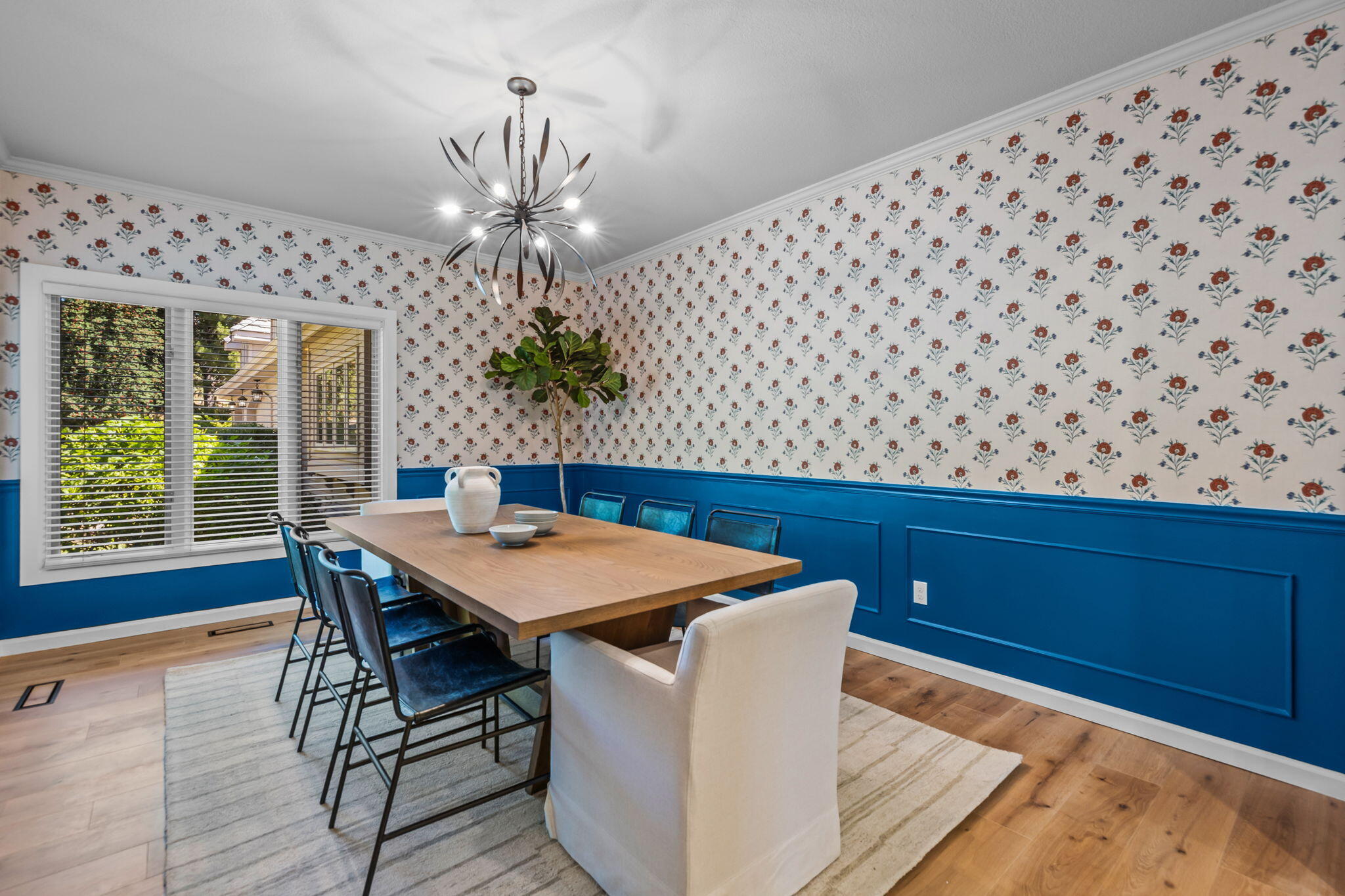 Dining room with blue paneled walls, floral wallpaper, and a long wooden table beneath a sculptural chandelier.