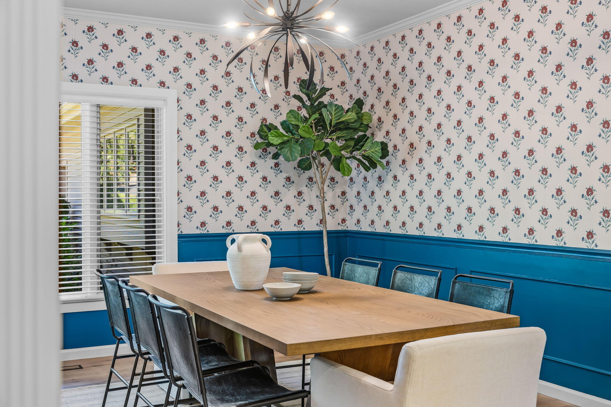 Dining room with a wood table, floral wallpaper, blue wainscoting, and sculptural chandelier.