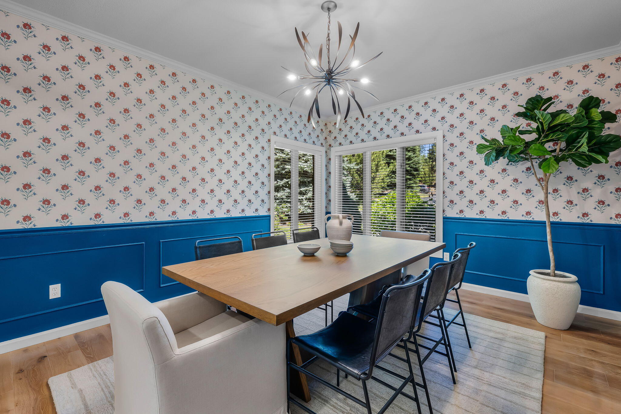 Dining room with blue paneled walls, floral wallpaper, a wood table, and sculptural chandelier.