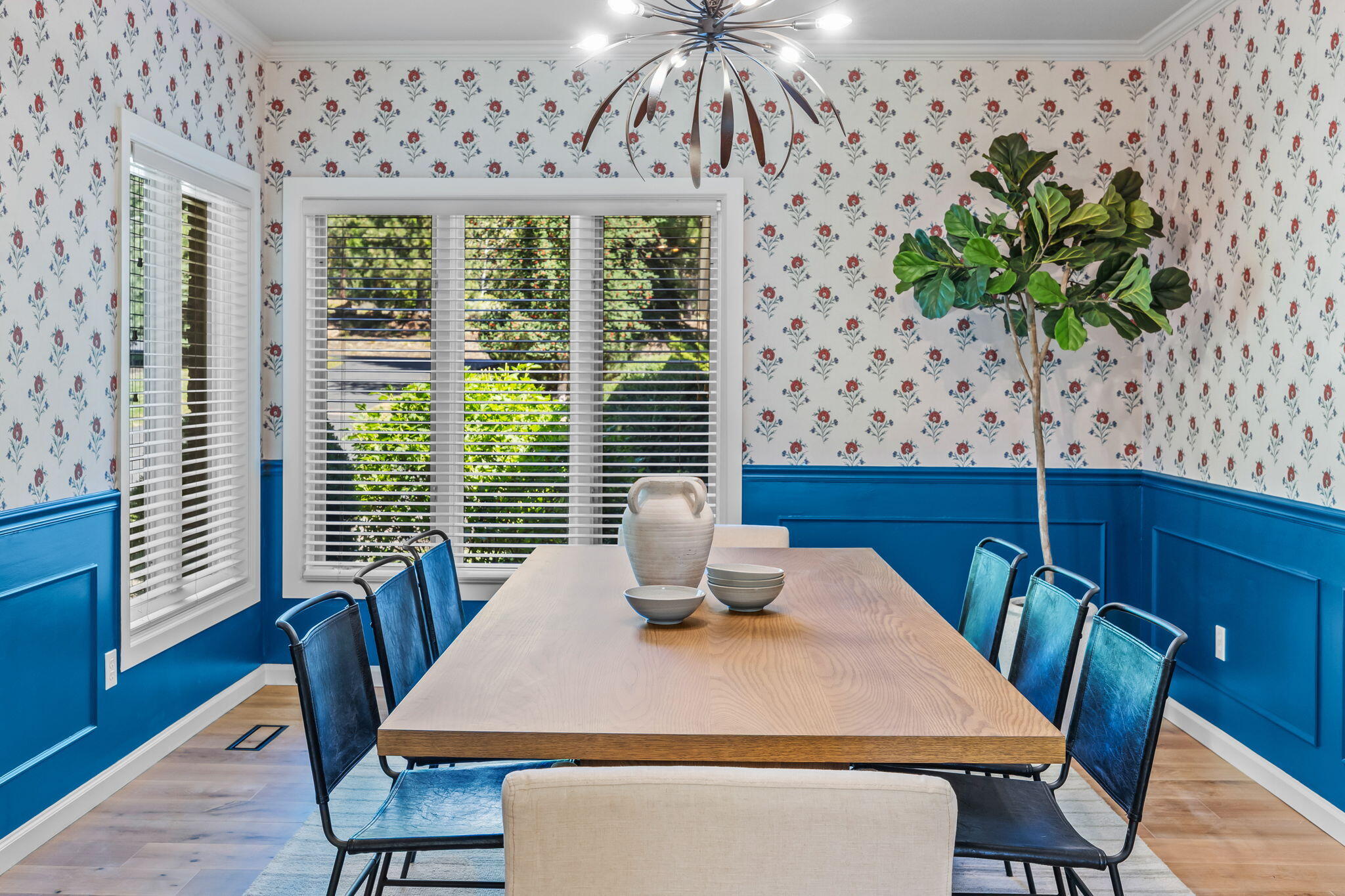 Dining room with a light-wood table, blue chairs, floral wallpaper, and bright blue wainscoting.