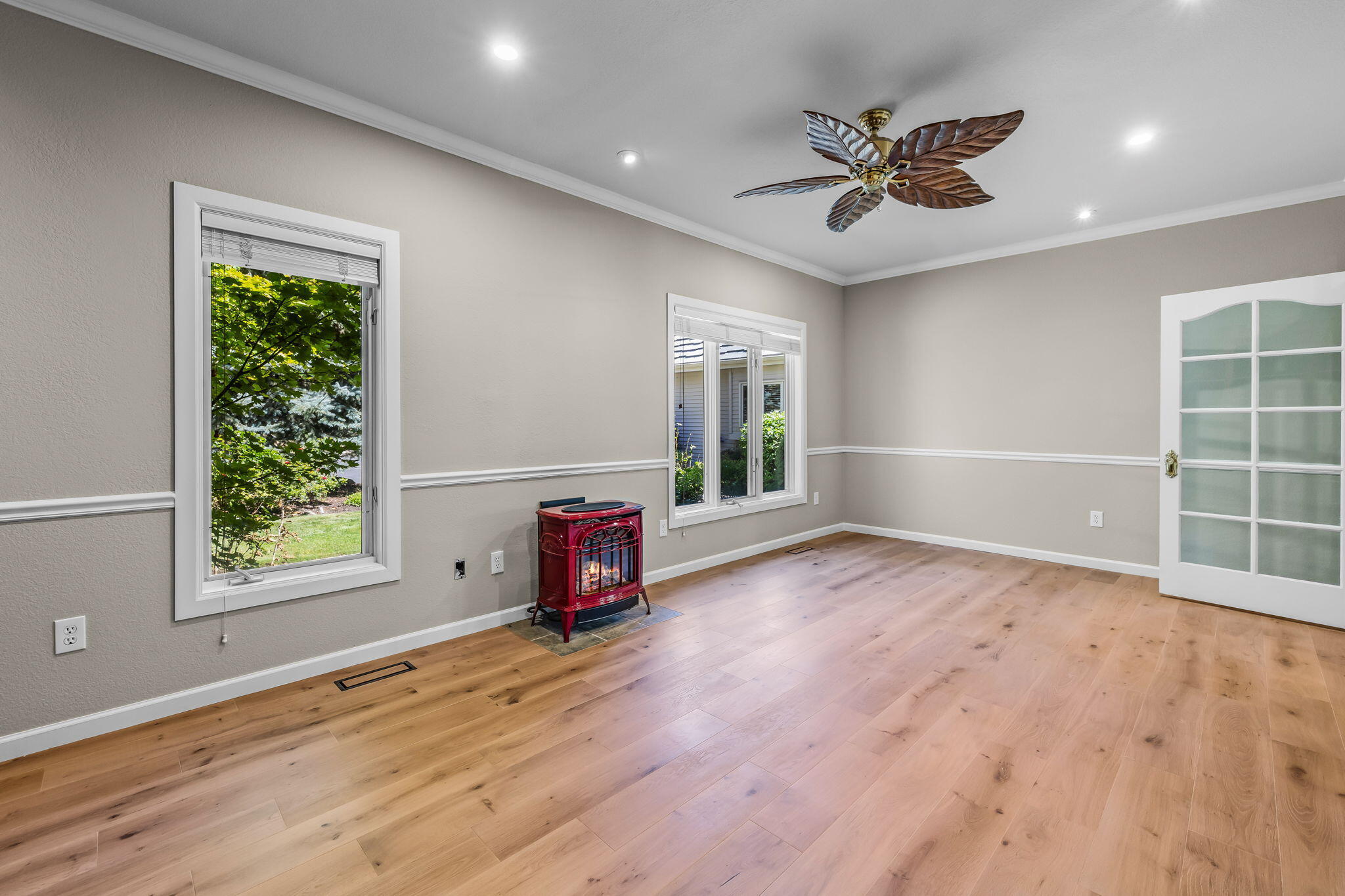 Empty room with light wood floors, a red freestanding stove, and windows overlooking greenery.