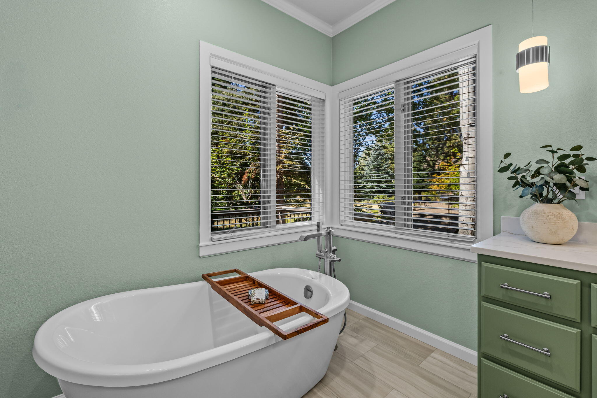 Bathroom with a freestanding tub beneath corner windows, sage-green walls, and a wooden bath tray.